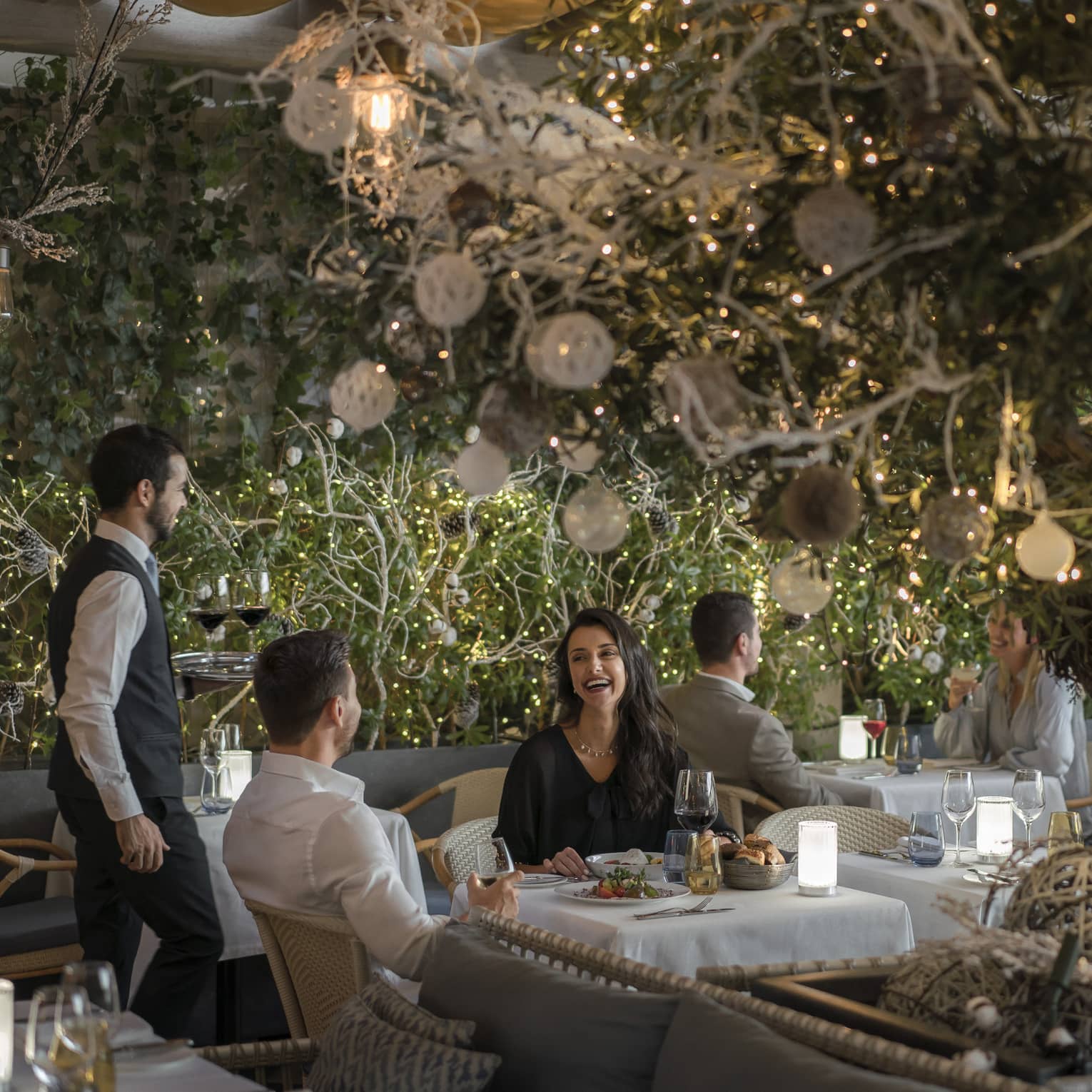 Male waiter wearing dark-grey pants and a vest over a white shirt attends a couple sitting at a white-clothed table set beneath a large tree adorned with white and silver Christmas decorations