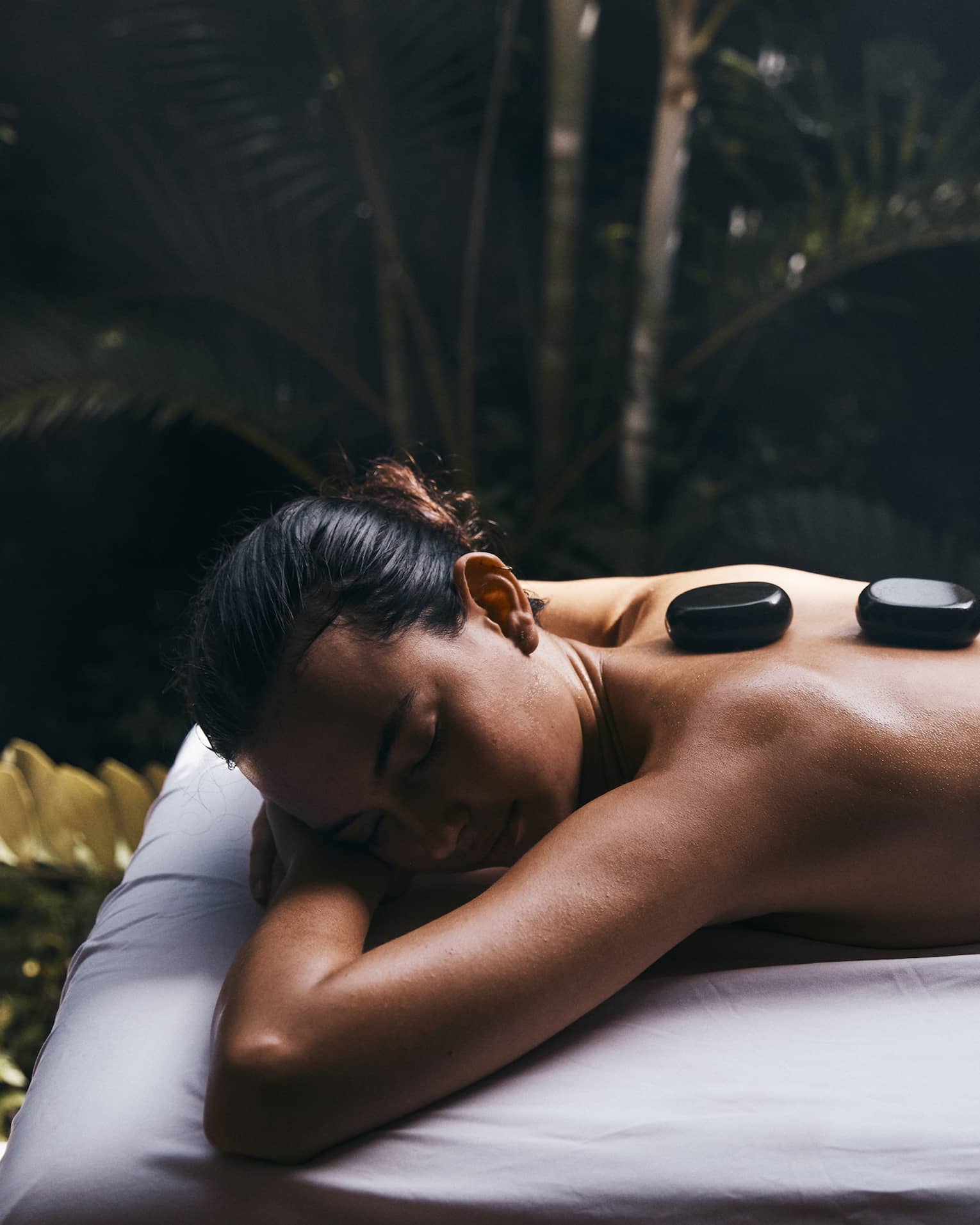 A guest on an outdoor massage table relaxes on their stomach with four glossy black warming stones on their back.