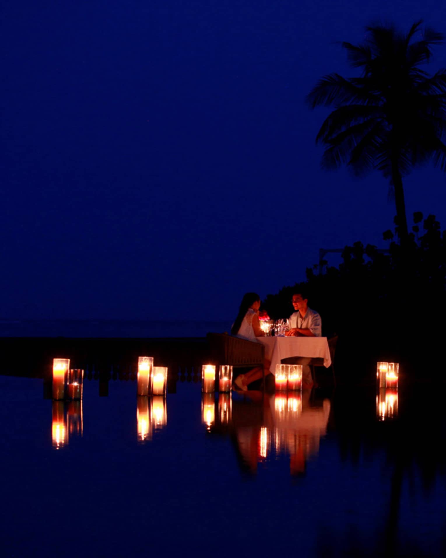 Couple dine by lagoon at small table surrounded by glowing candles at night