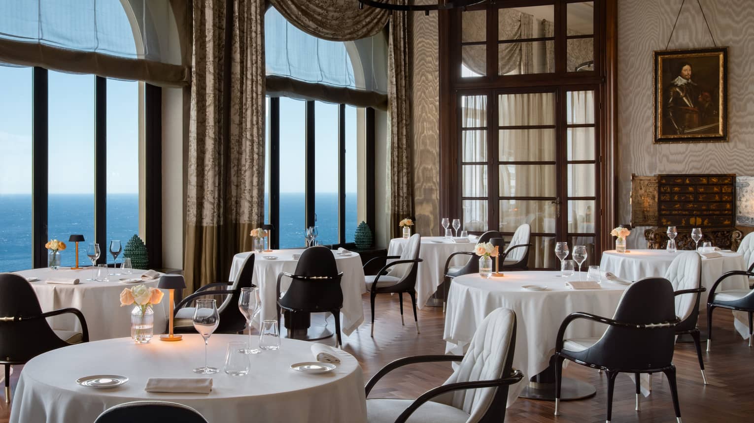 Indoor restaurant with white tablecloths, large drapery framing windows that look out to sea
