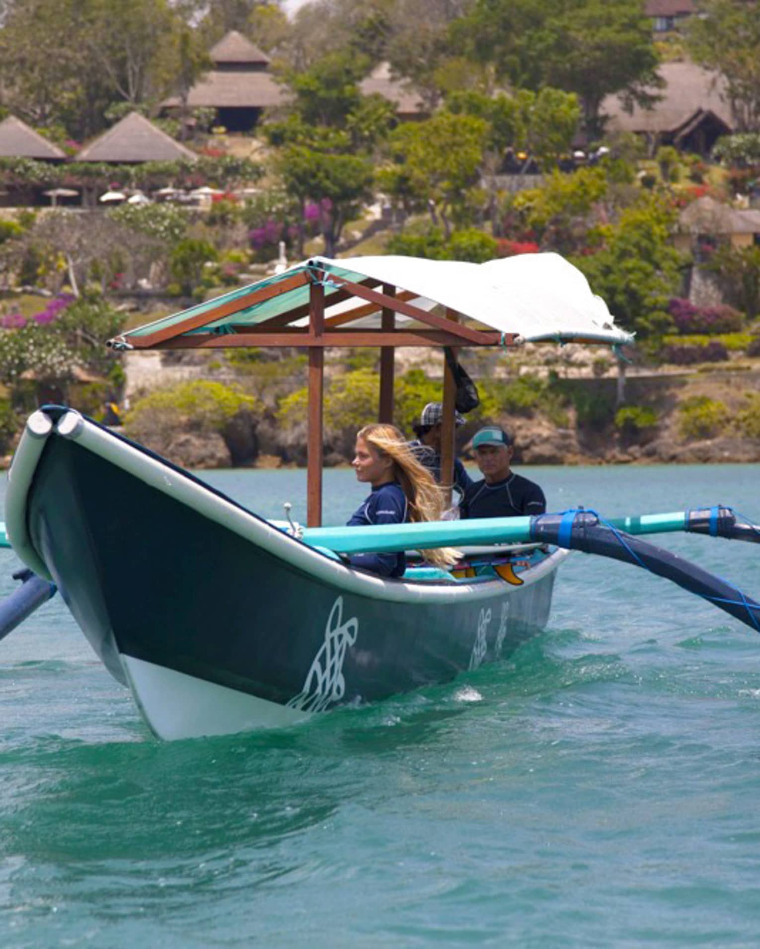 A guide sits at the stern of a covered outrigger canoe as two guests gaze at the water; behind, trees and huts dot the shore.