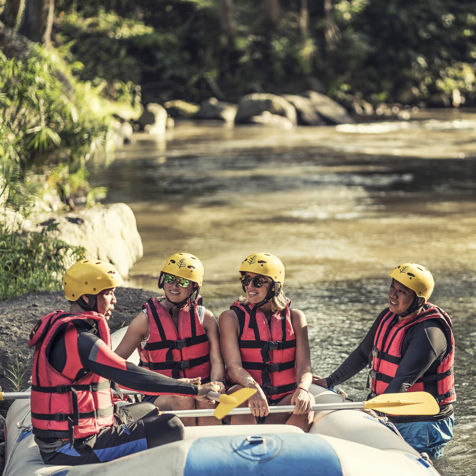 Four people wearing red life jackets, sunglasses and yellow helmets sit facing each other in a river raft