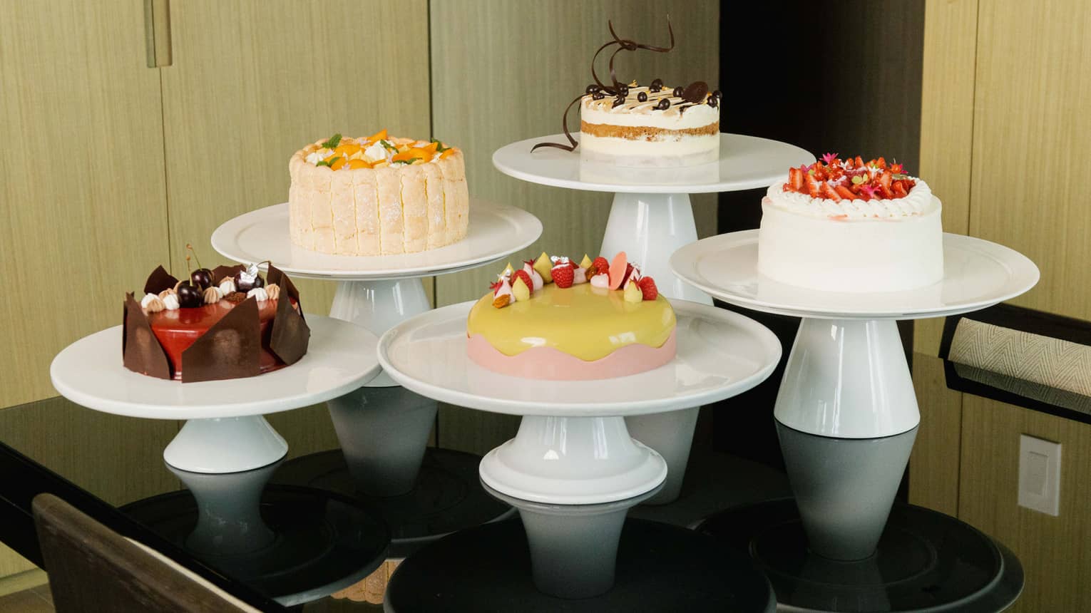 Collection of five cakes, each set on a white cake stand and gathered together on a black table