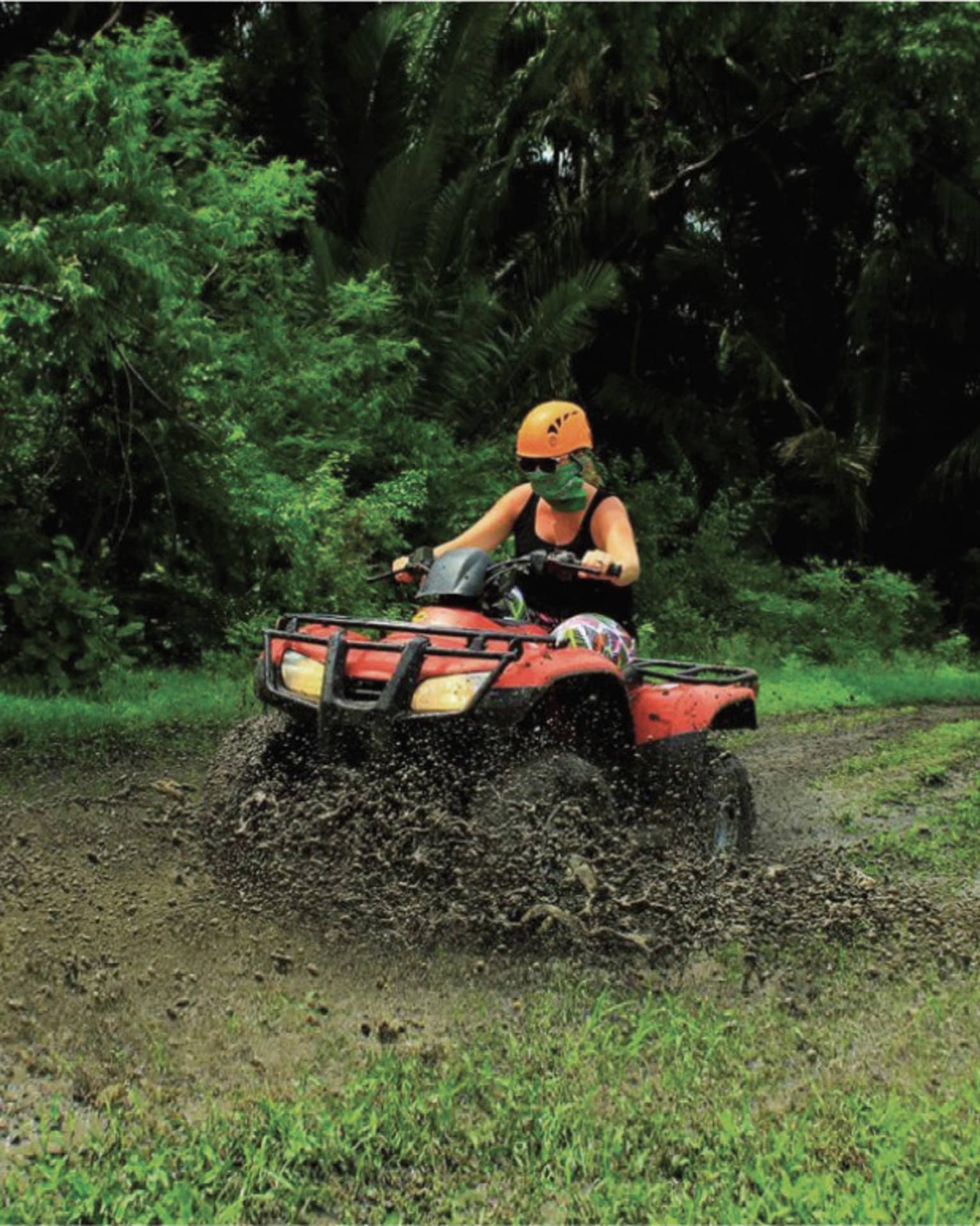 Person wearing a black tank top and bright orange helmet rides through the mud on a red ATV