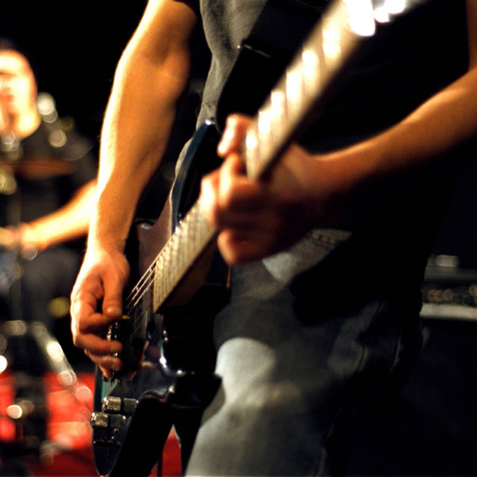 Close-up of musician's arms and hands playing an electric guitar with drumset in the background.