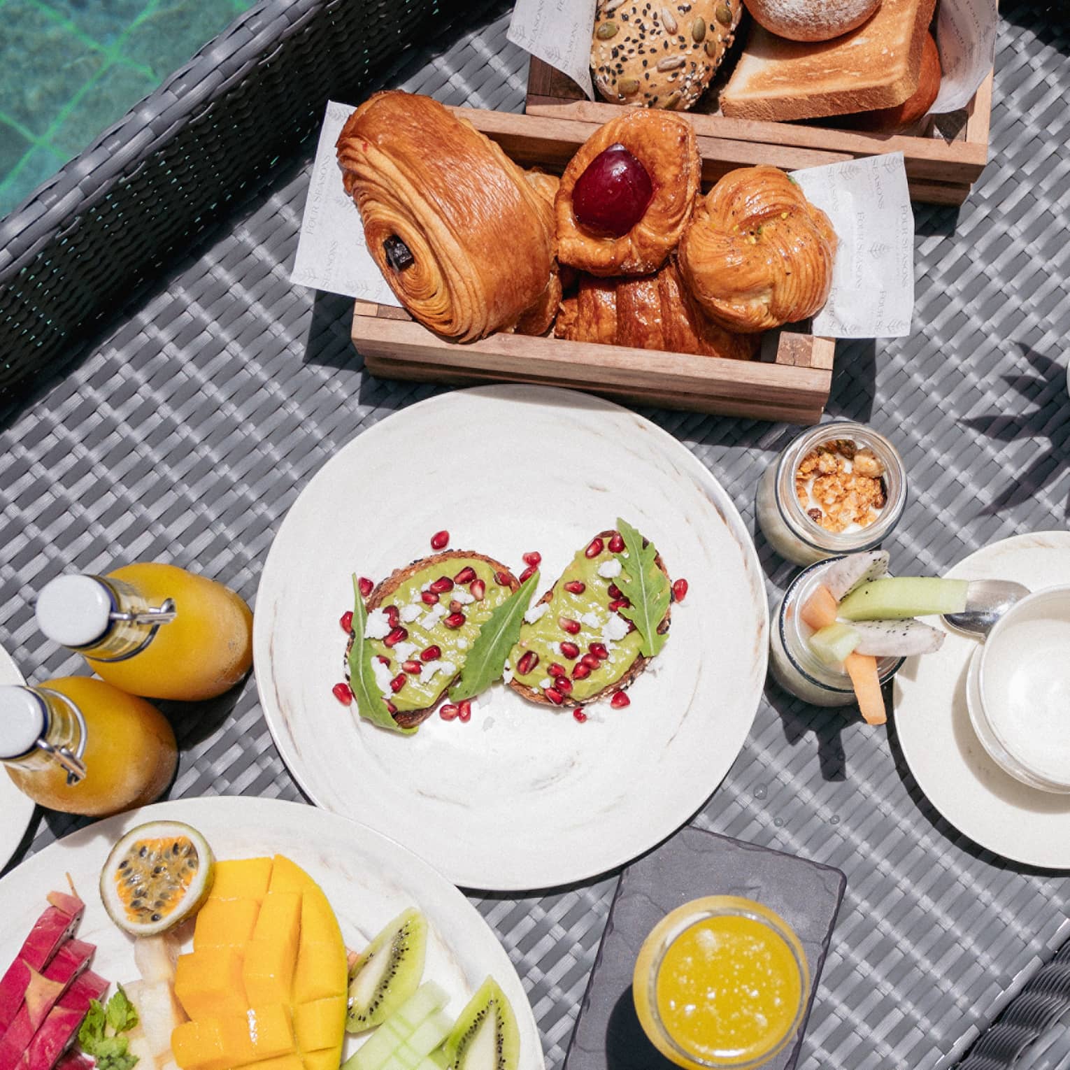 Close-up view from above of a wicker tray filled with fruit, pastries, juice and toast floating in a crystal-clear pool.