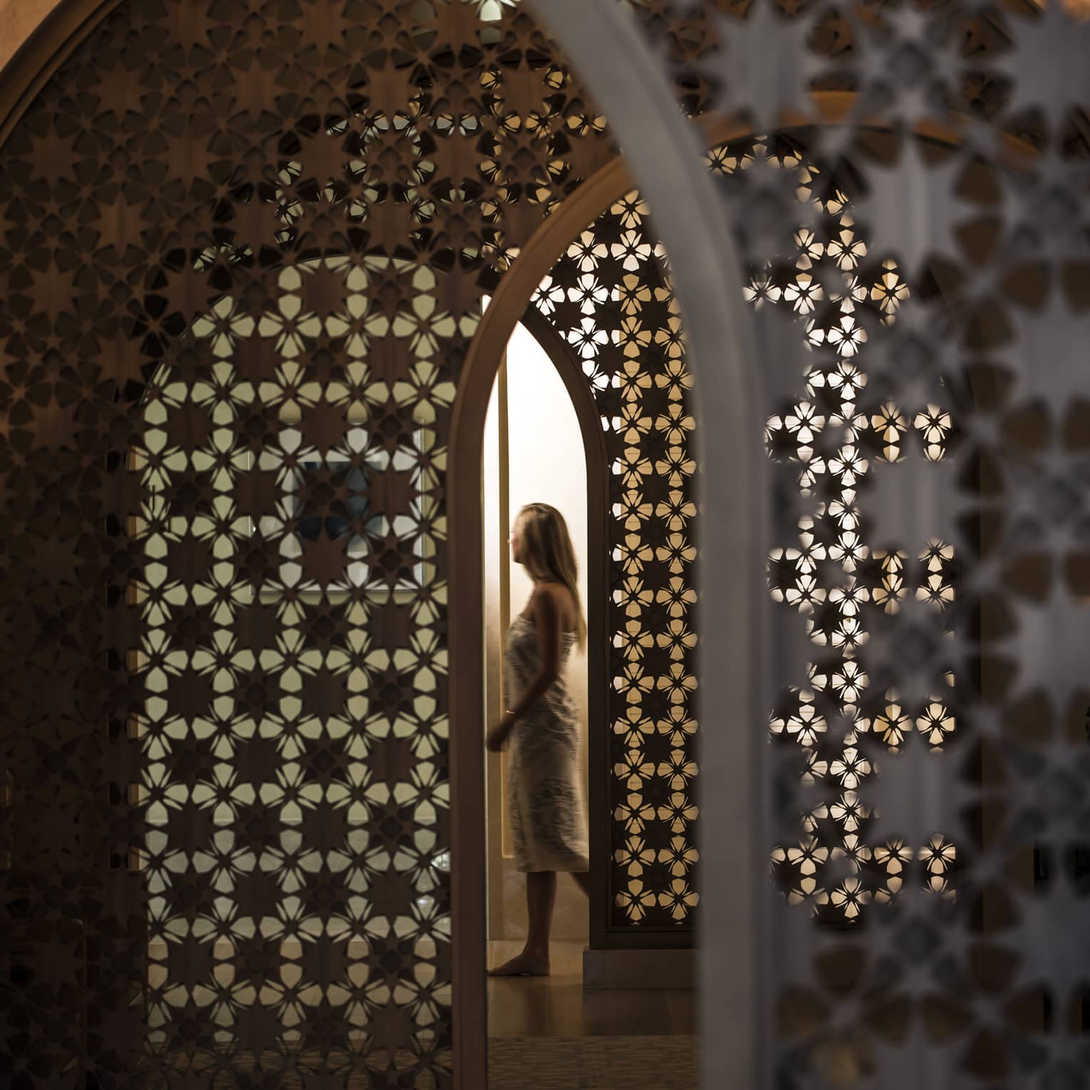 Woman in white wrap walks through dimly-lit decorative entranceway in hall