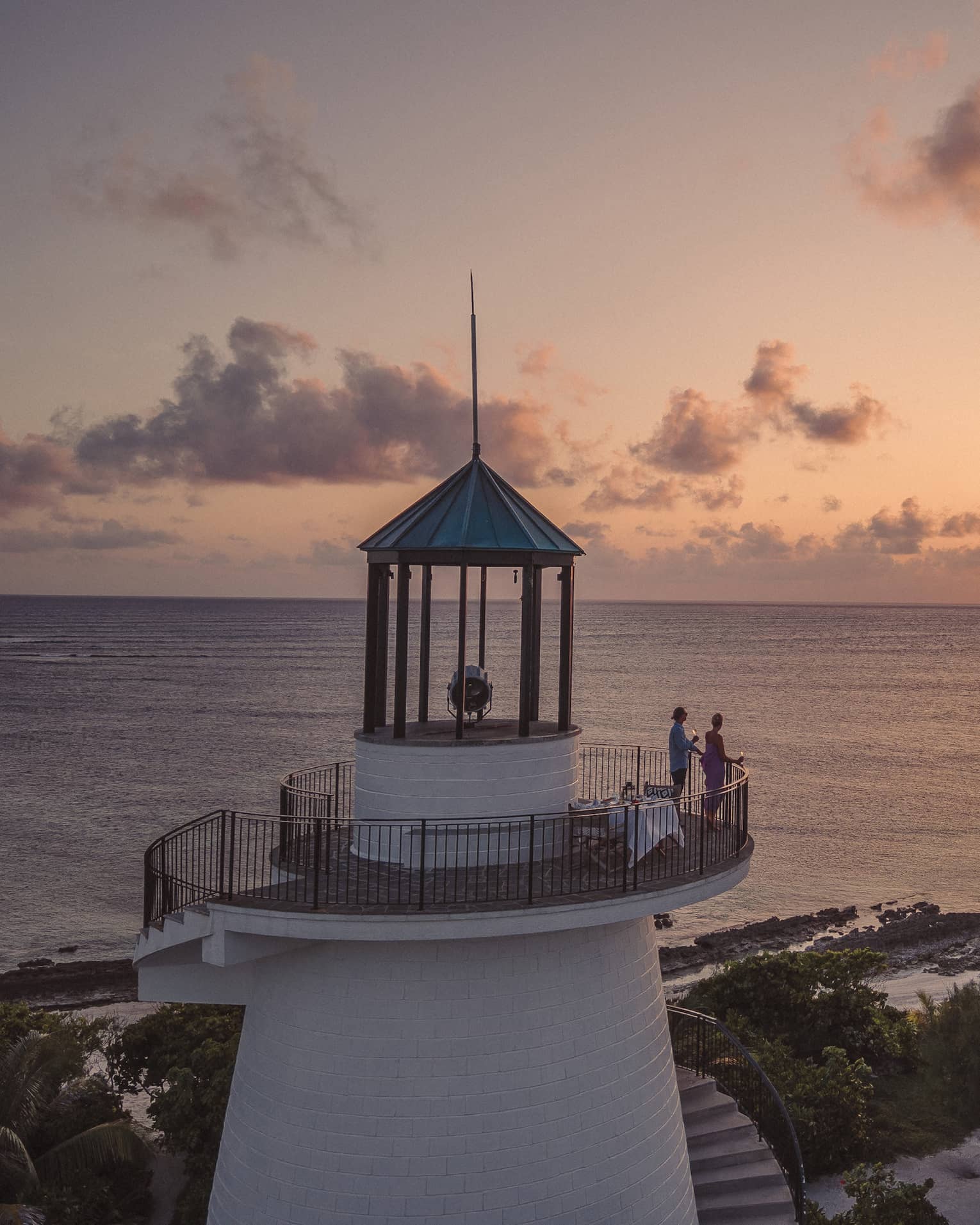 Long view of two people on a beach lighthouse balcony, enjoying the sunset over the ocean, distant silhouette of birds. 