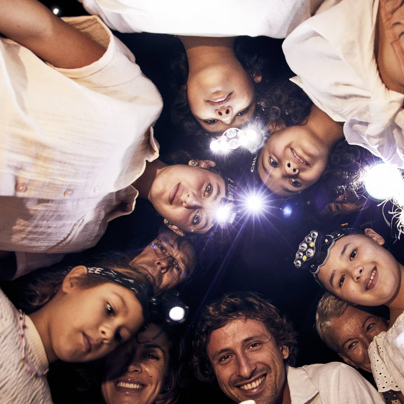 A group of children and adults, some wearing headlamps, stand in a circle and smile downwards into the camera.