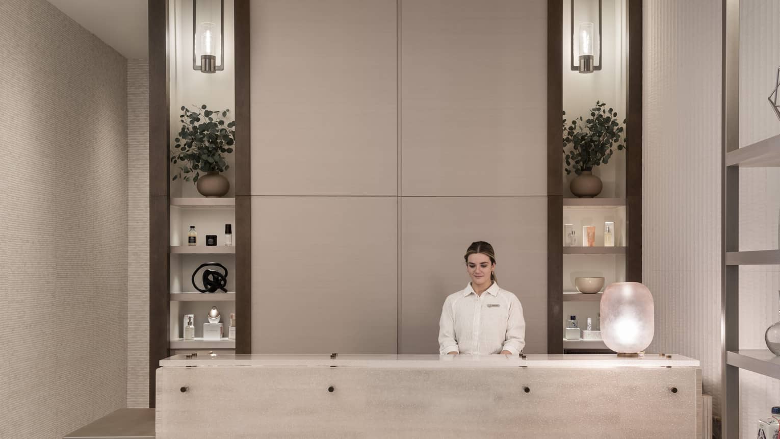 Receptionist in white shirt stands behind white marble spa reception desk with shelving display in backdrop