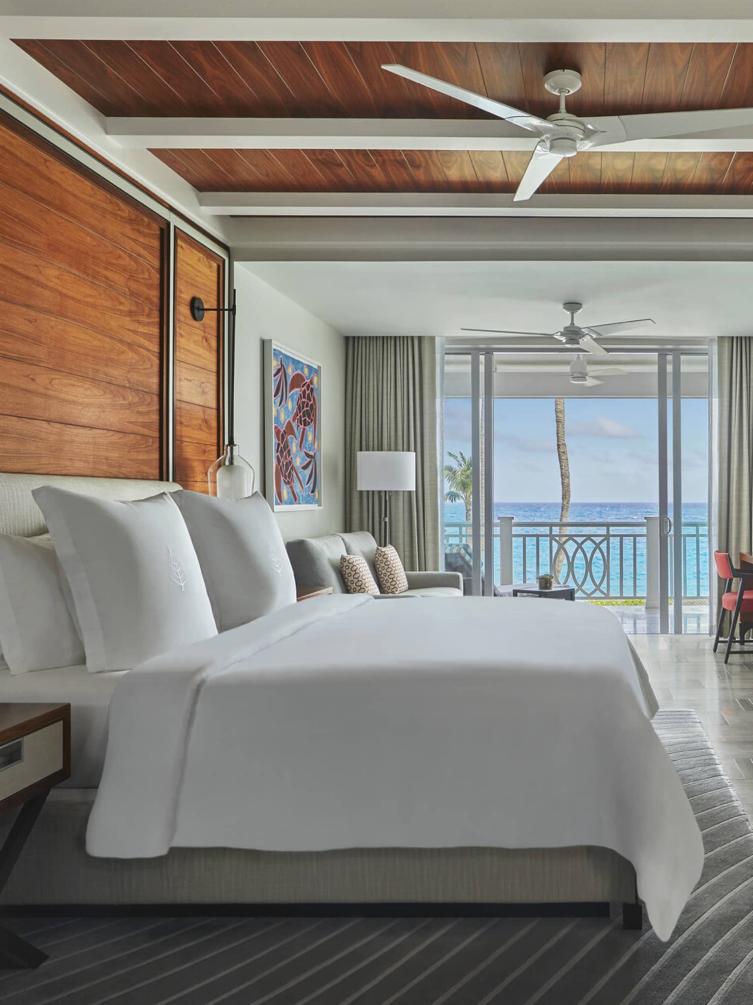 Ocean-view luxury resort bedroom with wooden accents, king bed, seating area, desk, wall-mounted TV and balcony