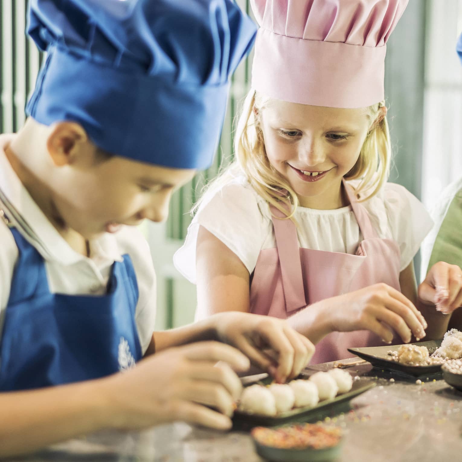 Three children in chef hats, aprons roll pastries at counter 