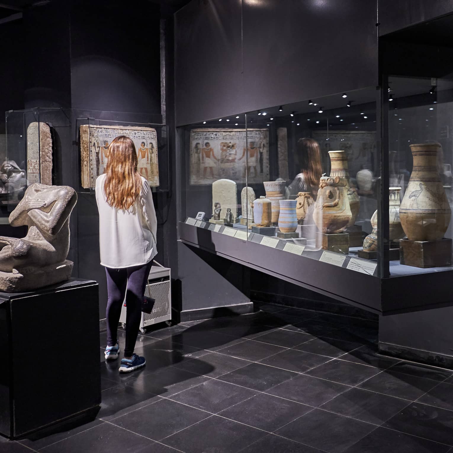 Rear view of a guest standing among artefacts in a dark museum; a glass case displaying stone jars creates a reflection.