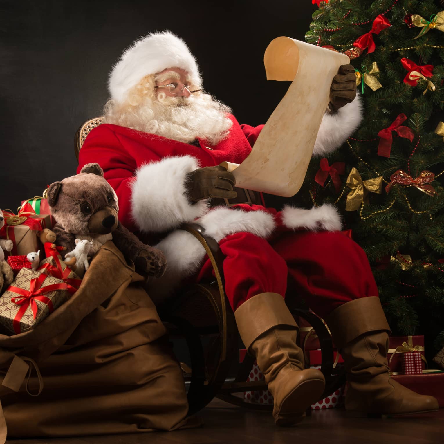 Santa Claus seated in large rocker holding a scroll, flanked by decorated Christmas tree with gifts and overflowing gift bag