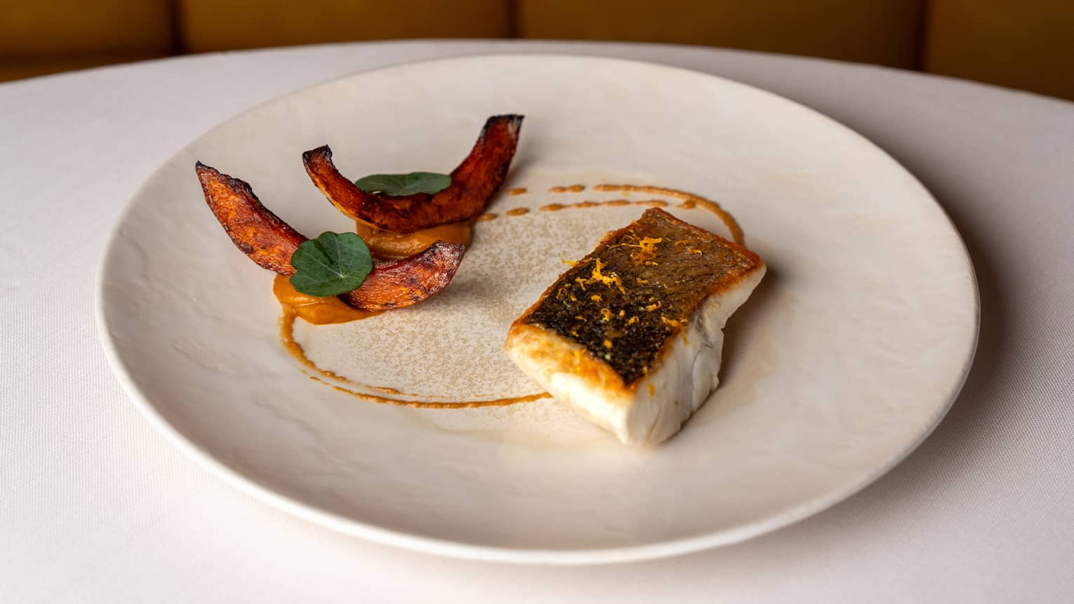 A round white plate holds a piece of white cooked fish, cooked slices of an orange-fleshed vegetable and a drizzle of sauce.