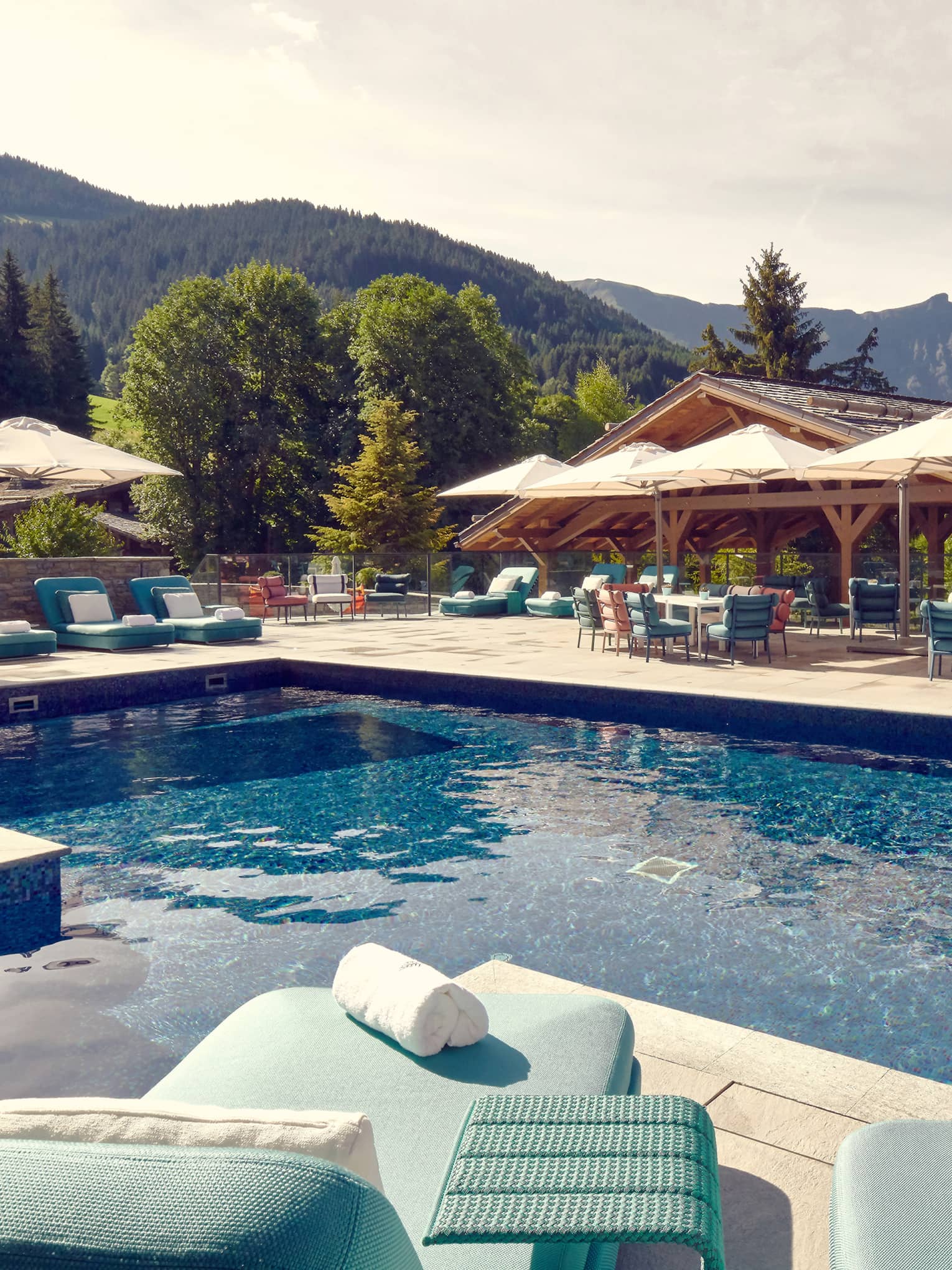 Outdoor pool with aqua blue chaises longues and beige umbrellas on surrounding deck and mountains in background