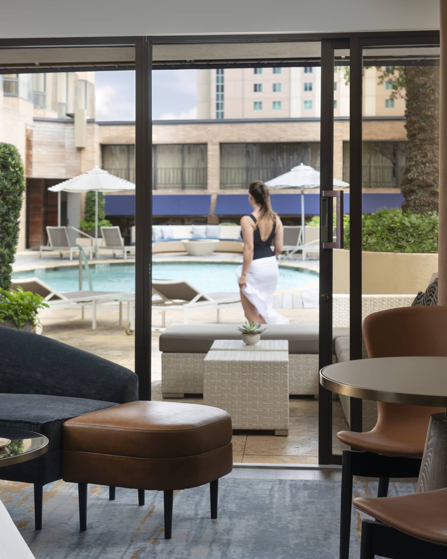 An indoor cabana decorated with modern furniture; outside the glass door a guest surveys the outdoor pool and lounge chairs.