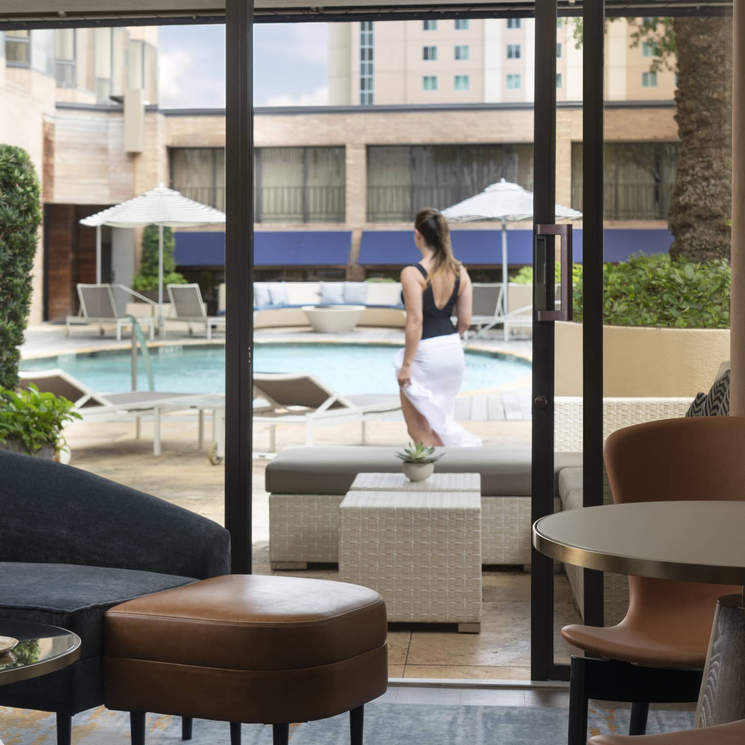An indoor cabana decorated with modern furniture; outside the glass door a guest surveys the outdoor pool and lounge chairs.