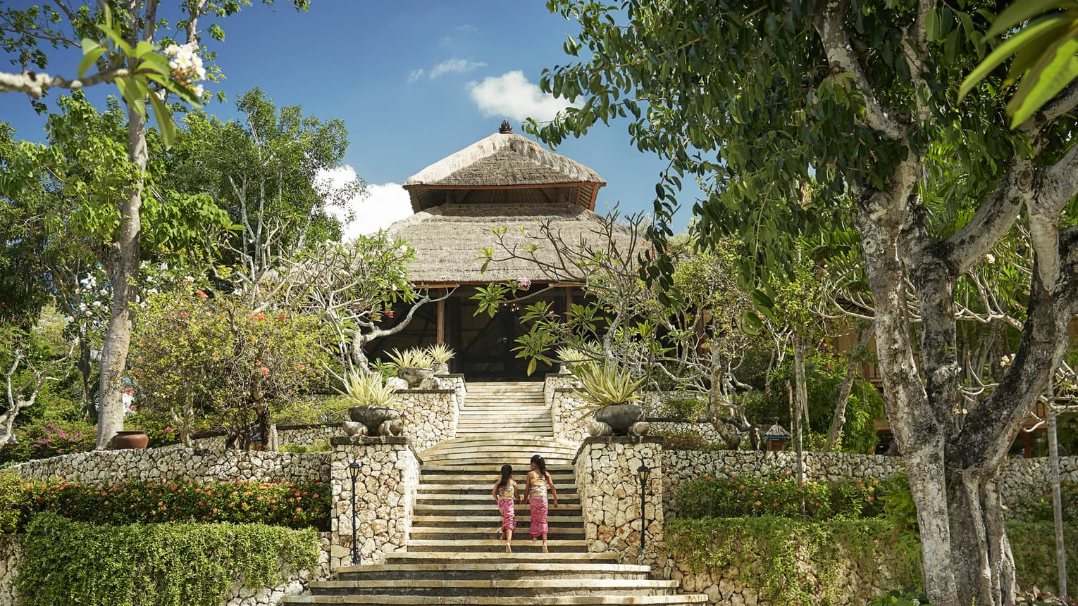 Back of two young girls walking up large stone staircase to stone lobby with thatched roof