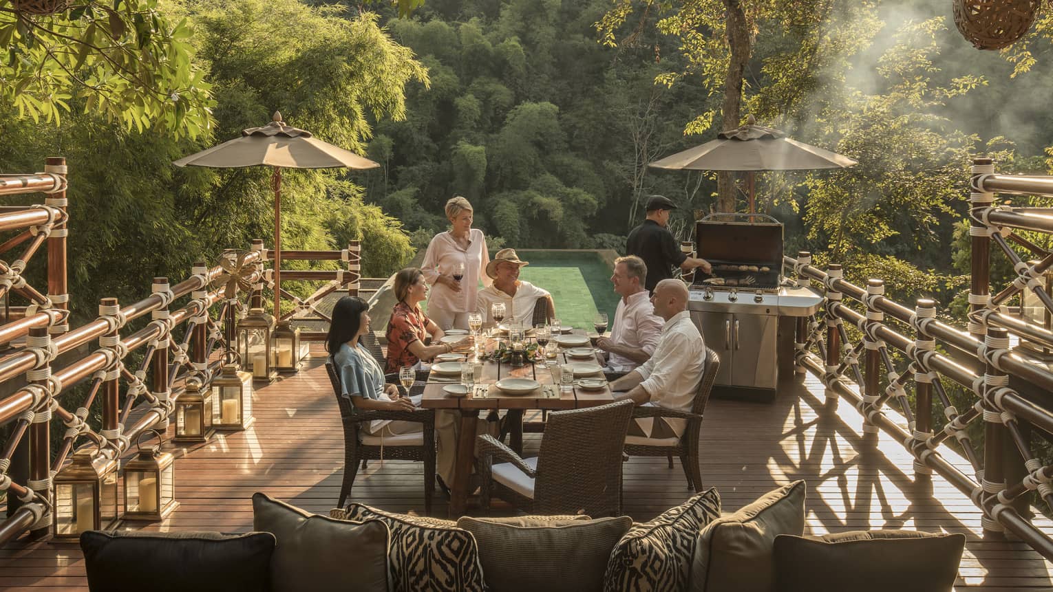 Smiling group of diners at patio table near chef at barbecue, surrounded by jungle