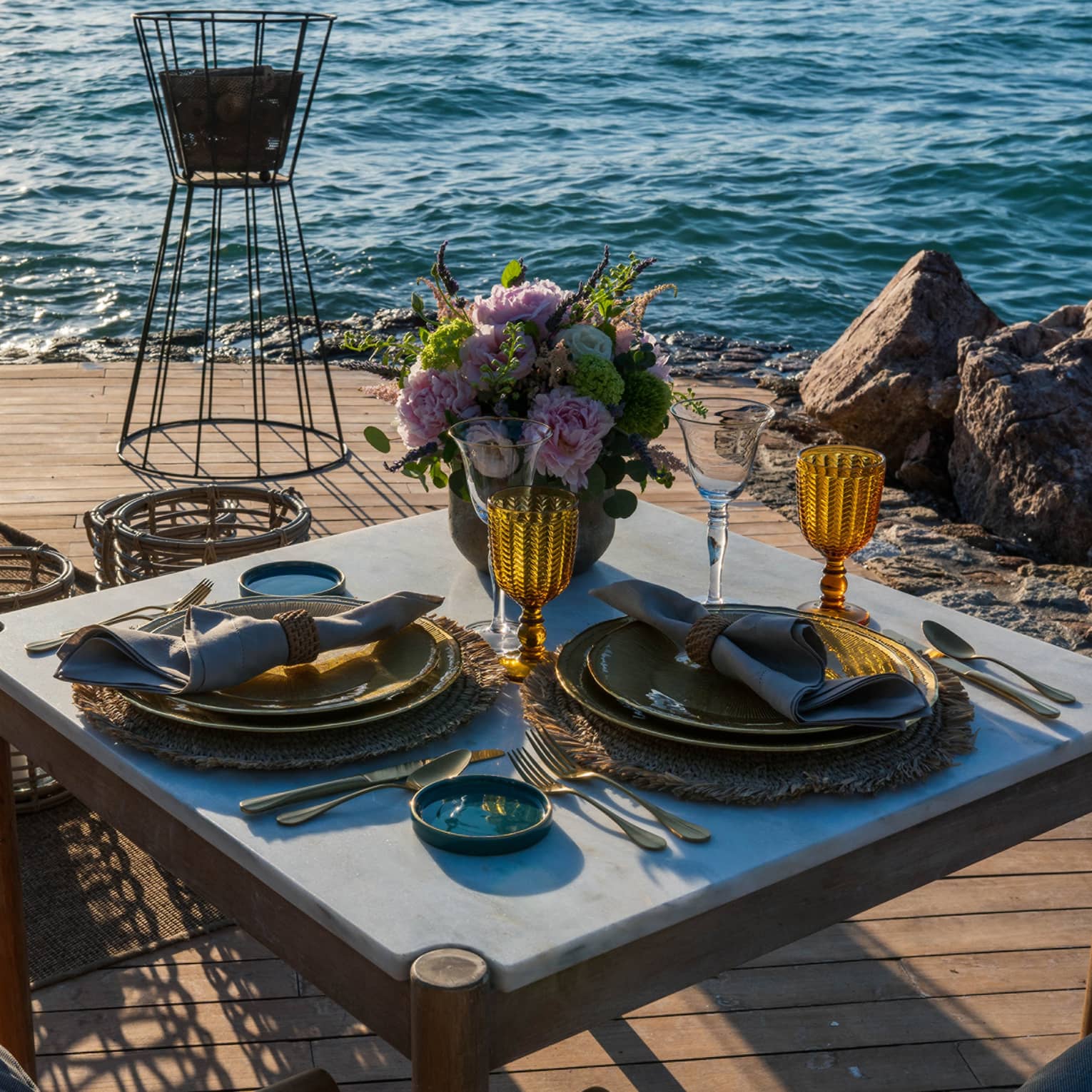 Elegant flowers and two rustic gold place settings with amber wine glasses grace a dining table on a waterfront patio.