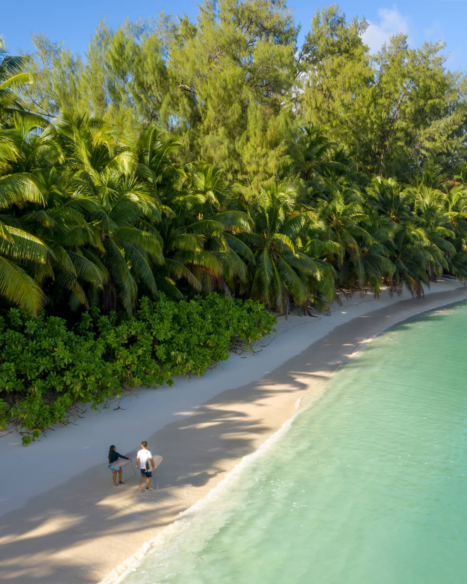 Couple walking along beach, carrying surfboards, flanked by lush tropical vegetation and blue-green water