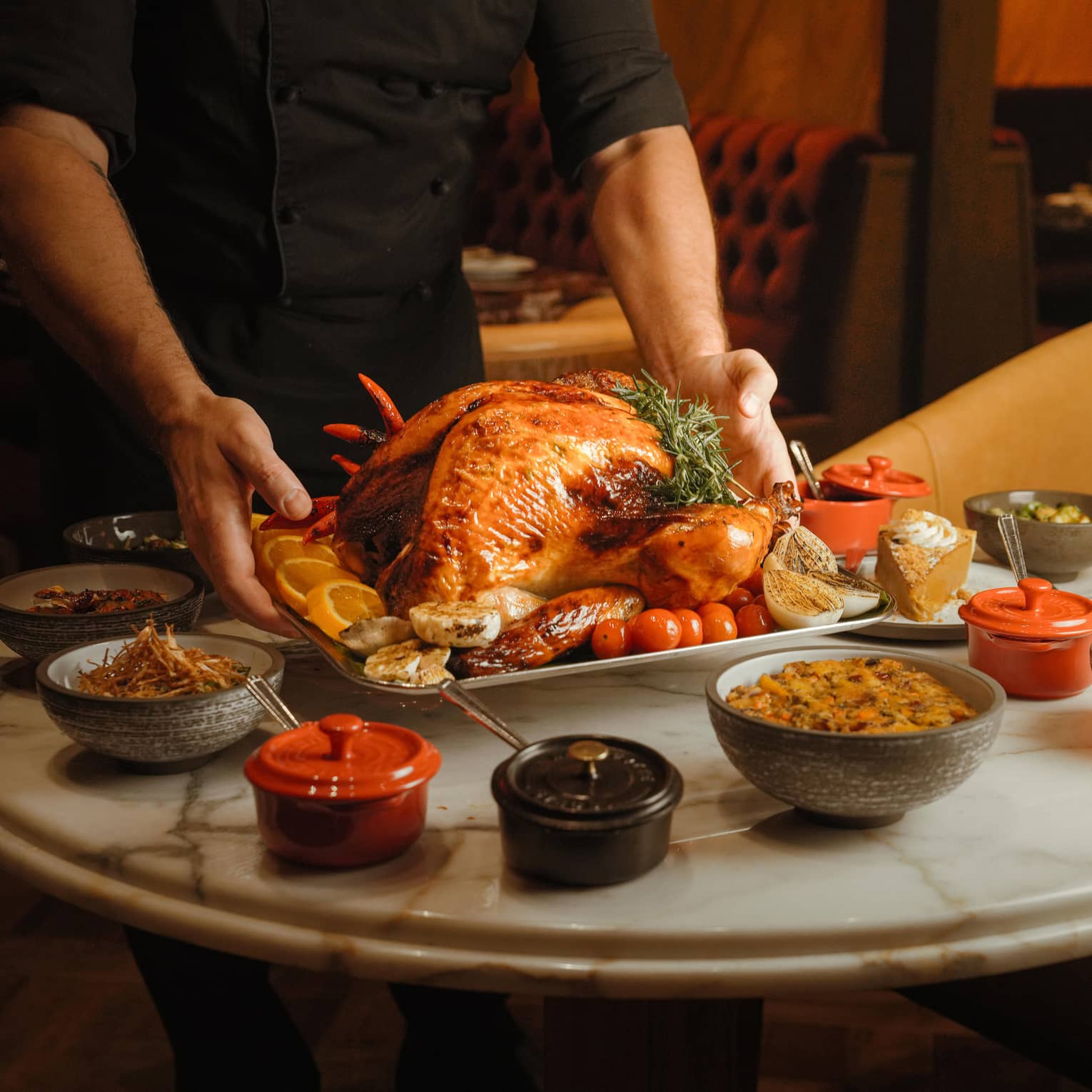 Server wearing black shirt places a whole roasted turkey on a white, round marble table also filled with small bowls of holiday dishes