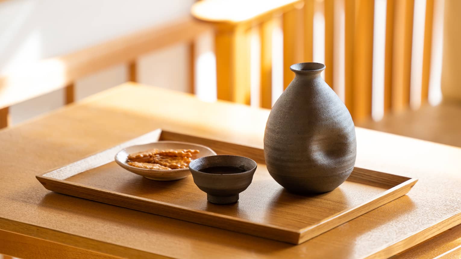 Close-up of a dark ceramic sake carafe with matching cup and a small plate of crackers on a wooden tray and table.