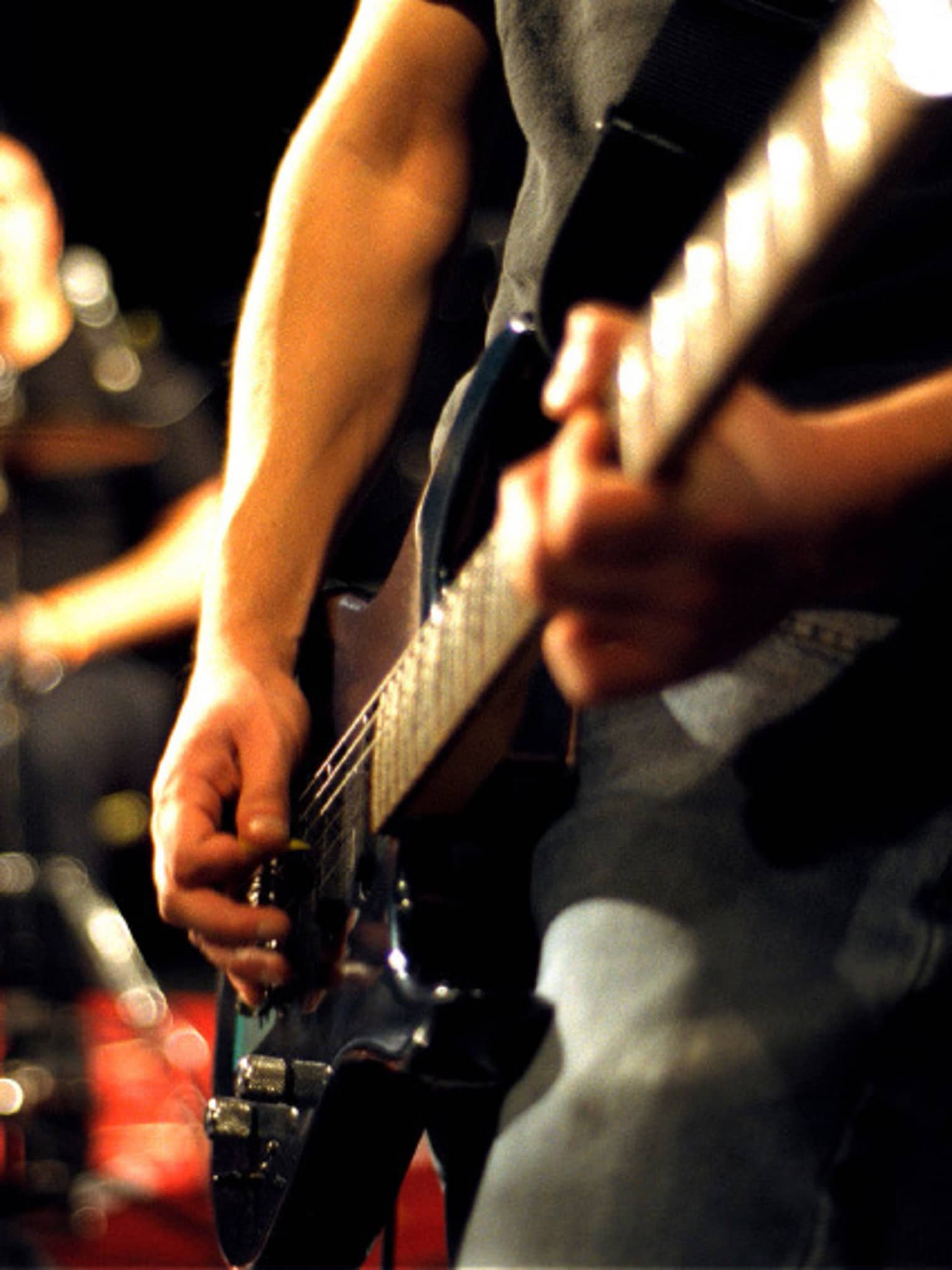 Close-up of musician's arms and hands playing an electric guitar with drumset in the background.