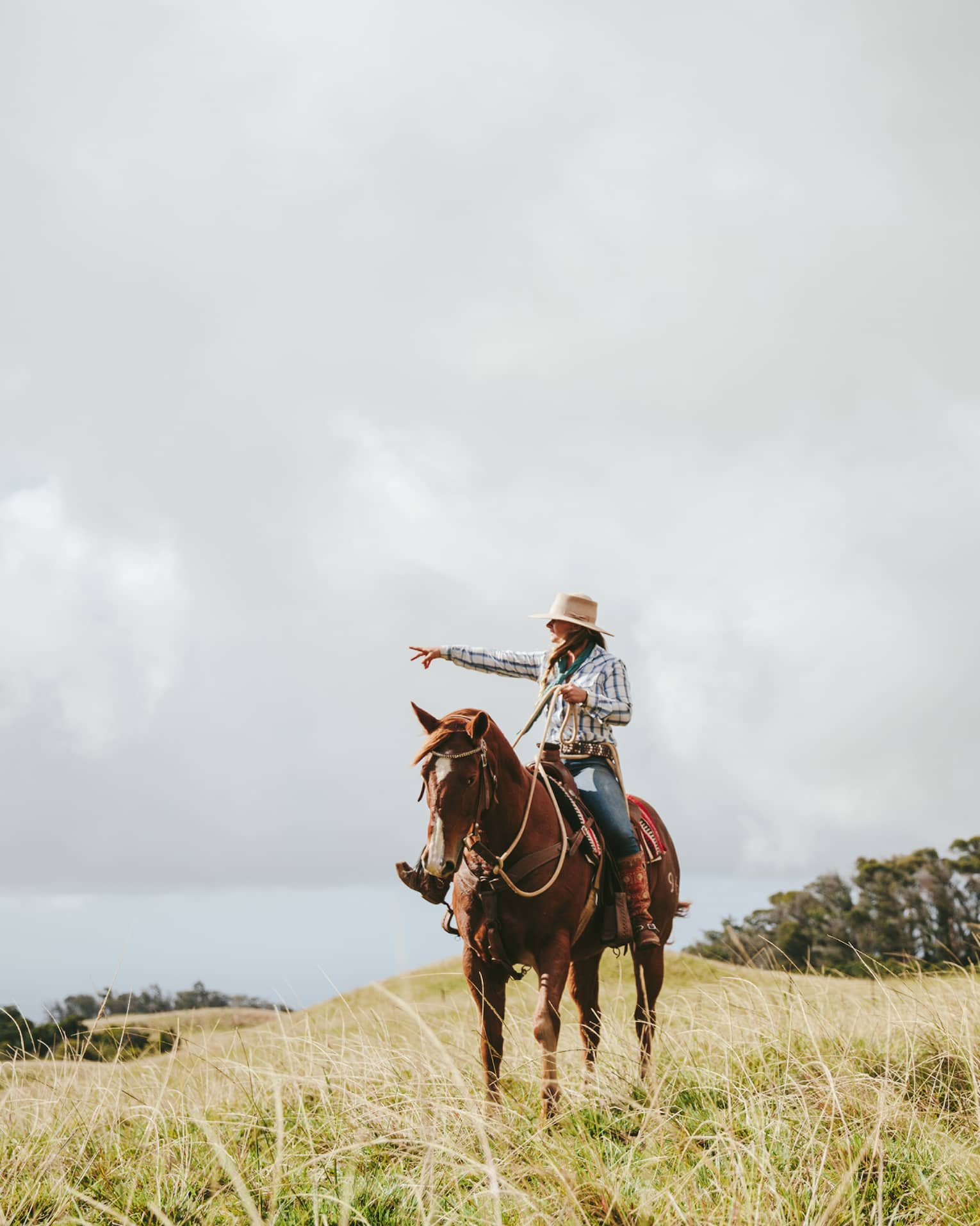 A couple riding horses, looking out at the long grass fields, with grassy hills and trees in the distance on a cloudy day.