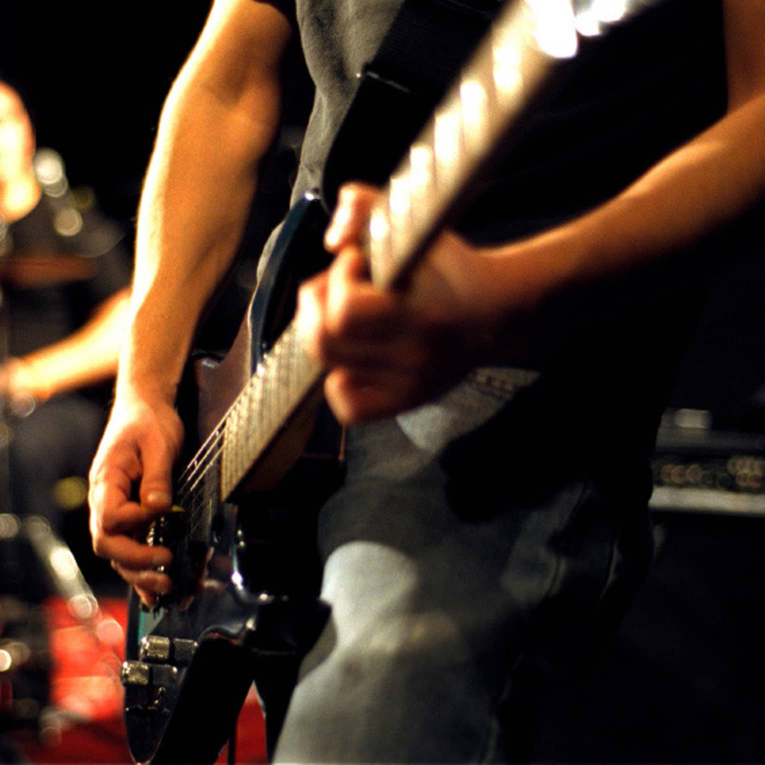 Close-up of musician's arms and hands playing an electric guitar with drumset in the background.