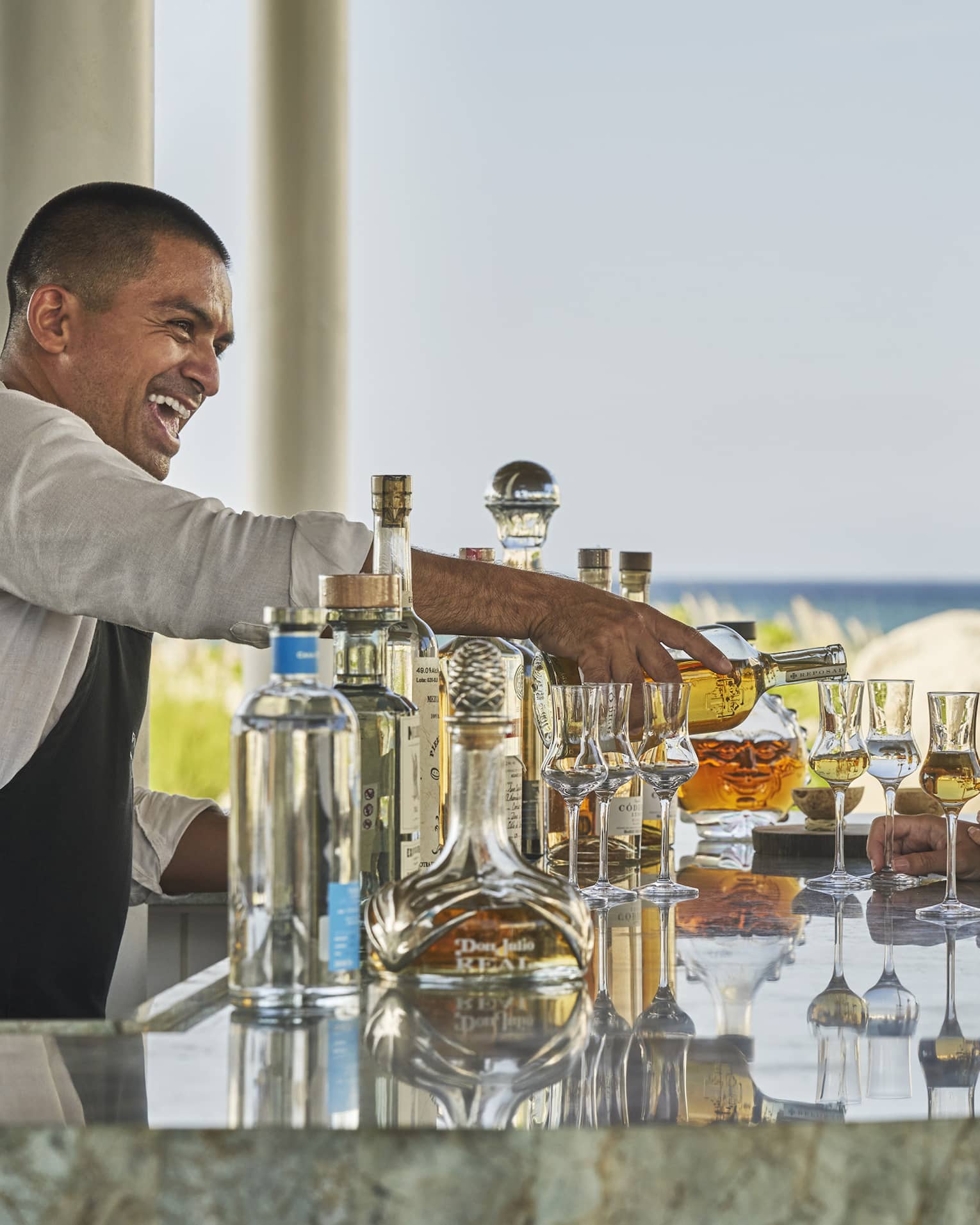 A male bartender serving drinks to a man and woman at a bar next to a beach.