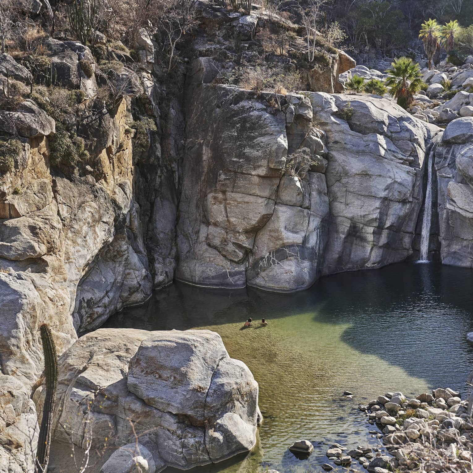Two people wade in a small ocean lagoon surrounded by large cliffs, creating a serene and picturesque scene with clear water and rugged natural beauty.