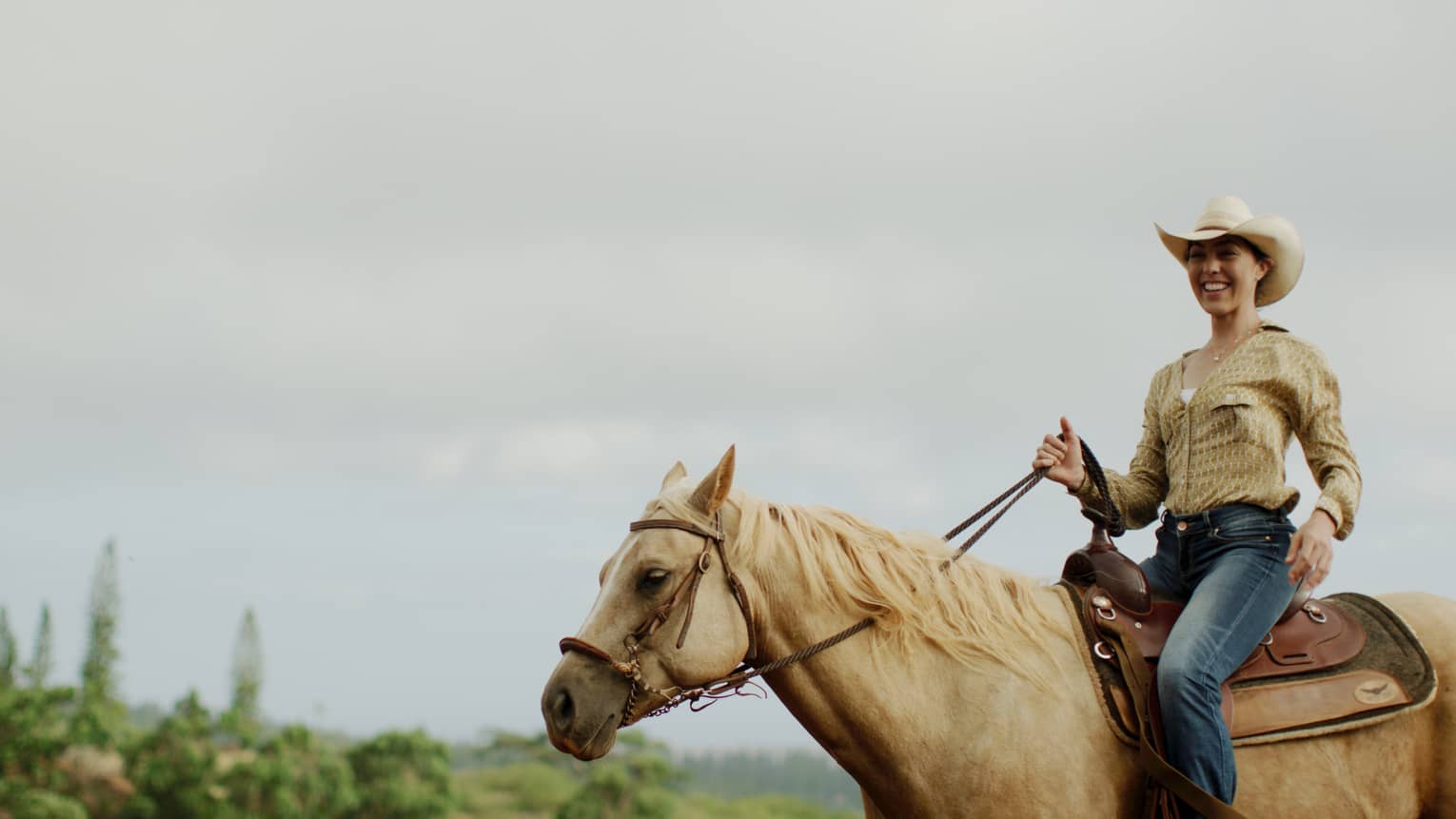 A smiling guest wearing a cowboy hat holds the reins while riding a tan horse under cloudy skies, forest in the distance.