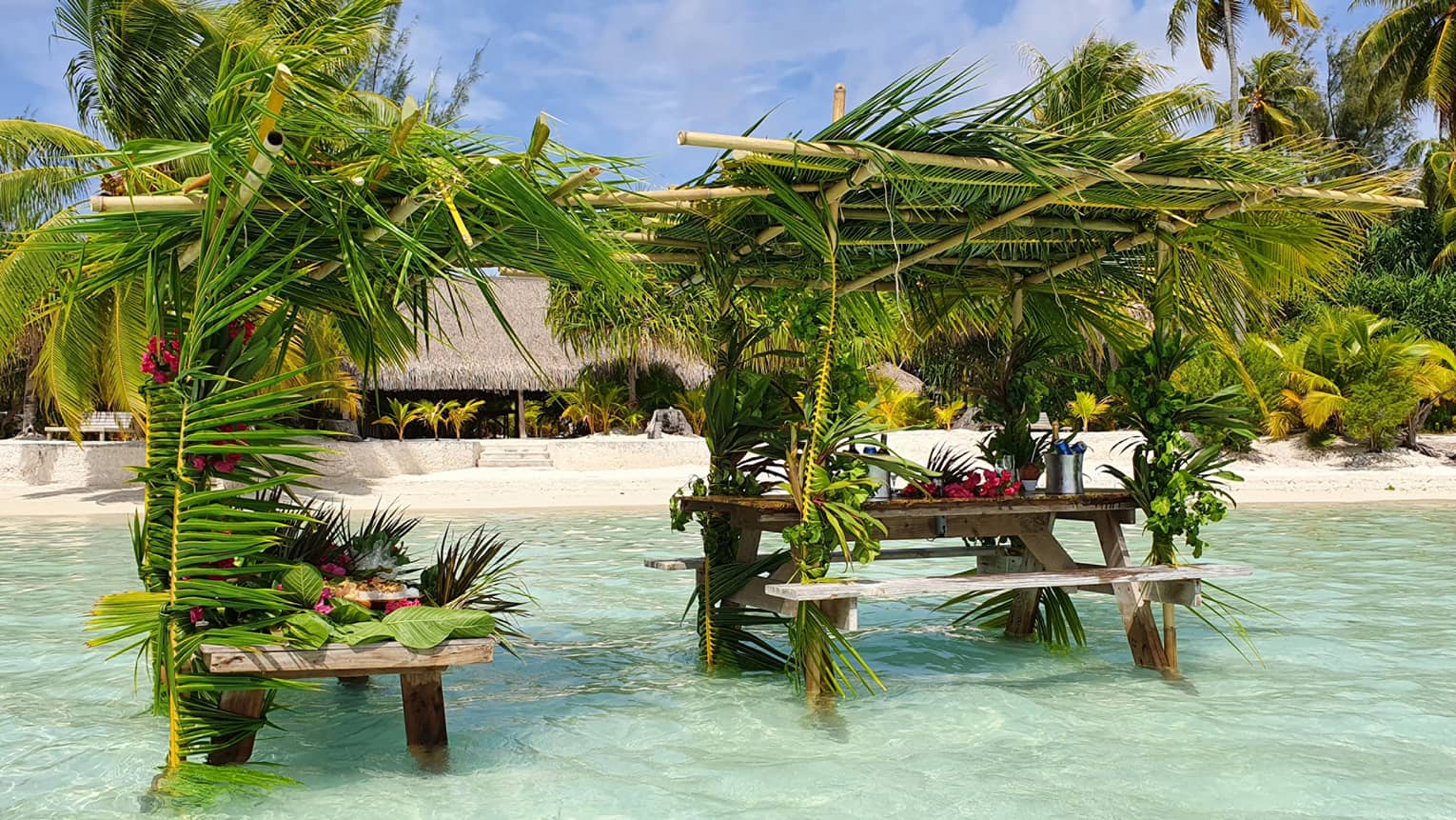 Picnic tables shaded by palm tree fronds sit knee-deep in clear turquoise water