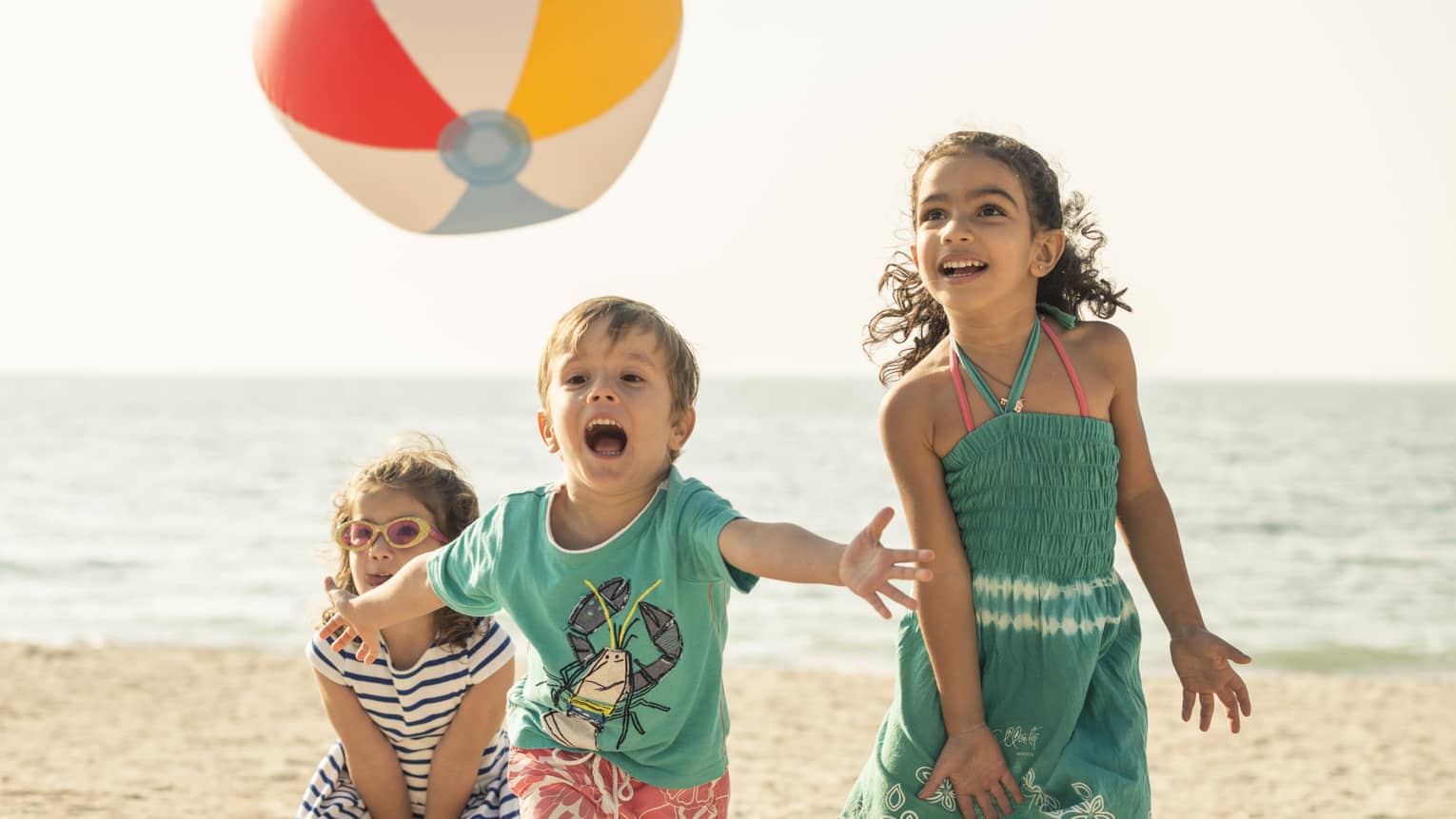 Close-up of three children playing with a beach ball on a sandy beach, the calm ocean and clear sky spanning behind them.