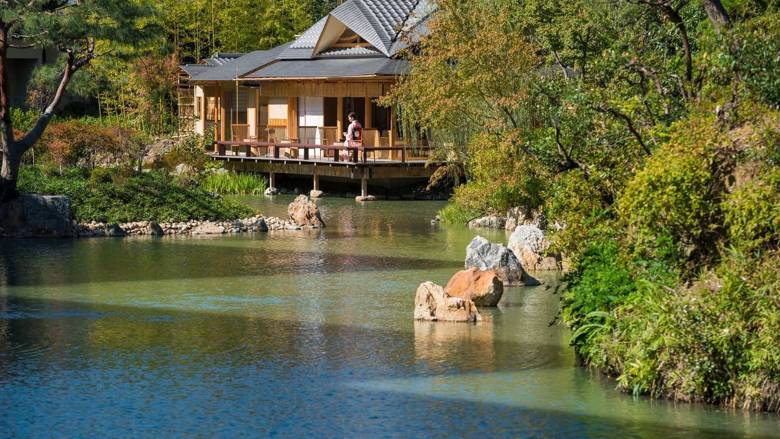 View across pond to bungalow where woman in robe sits on patio