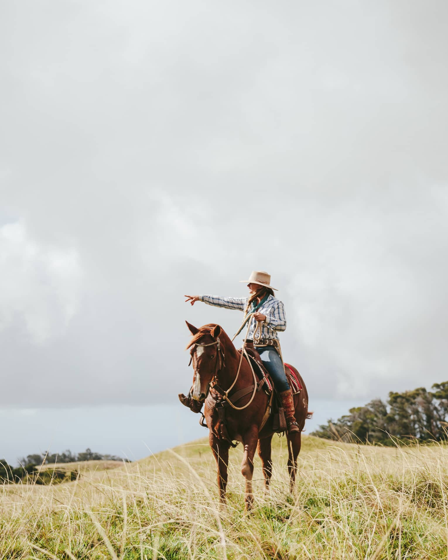 A couple riding horses, looking out at the long grass fields, with grassy hills and trees in the distance on a cloudy day.