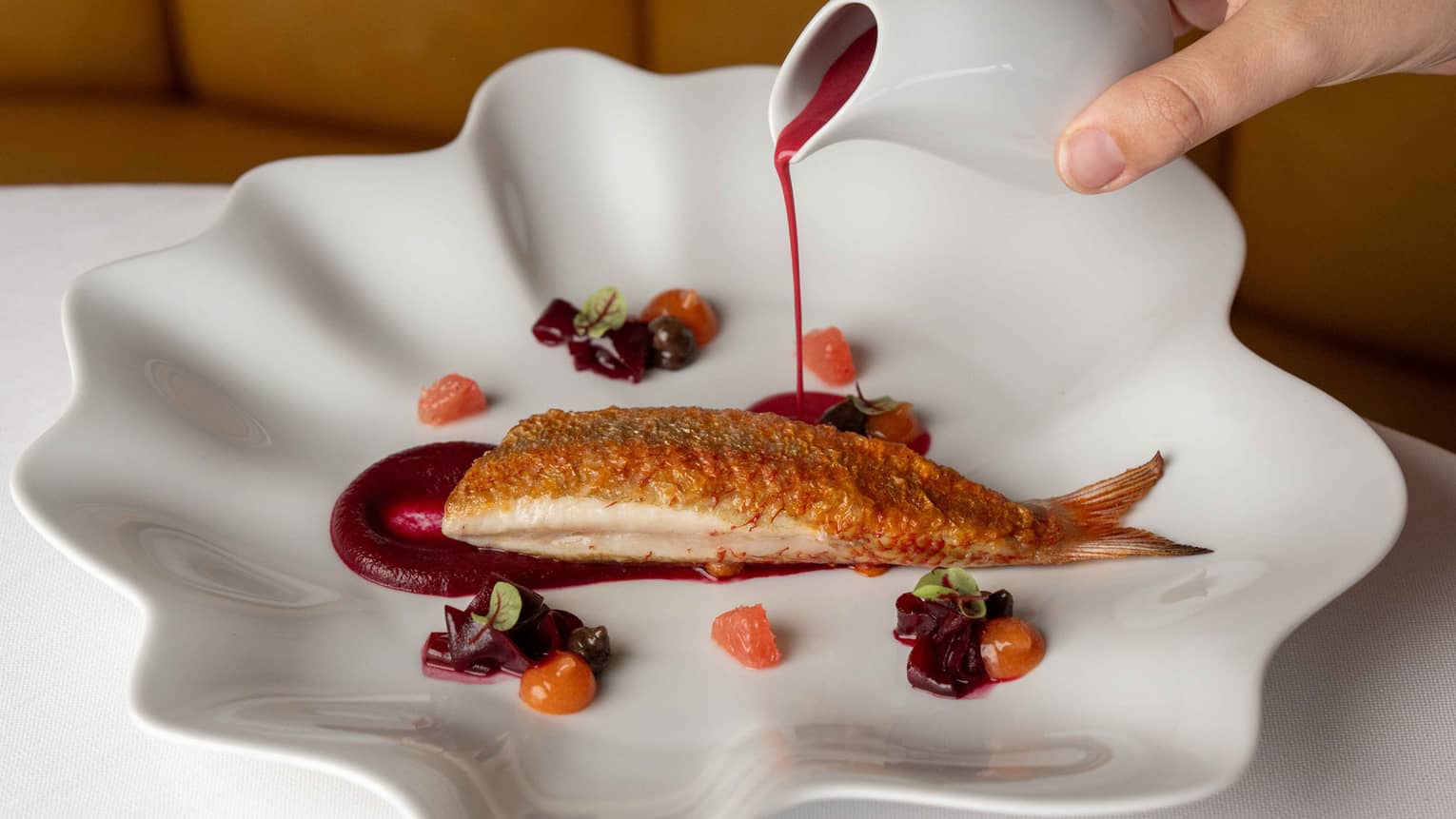 An artistic plate with a wavy edge holds a piece of cooked fish with garnishes and a sauce.