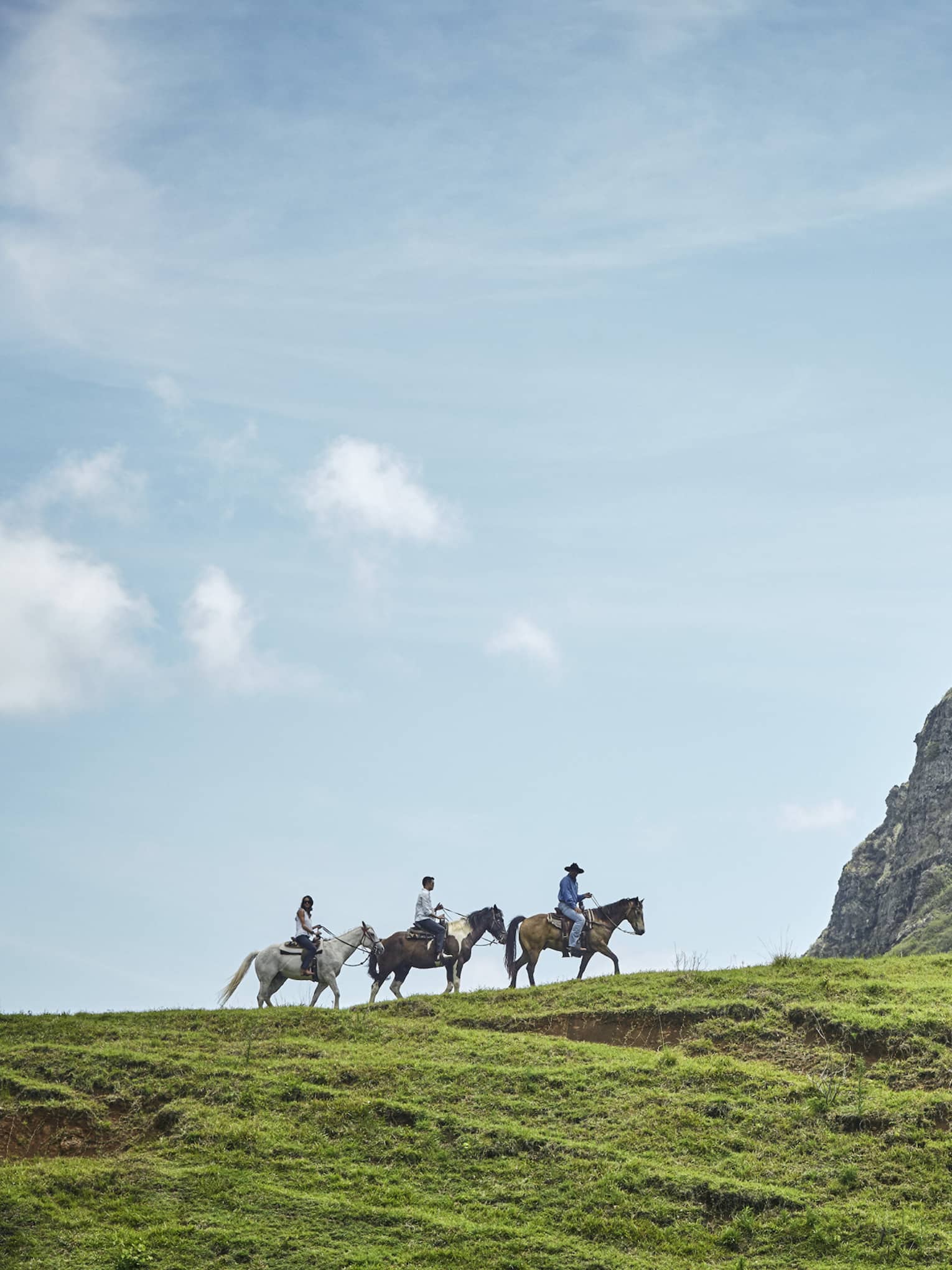 Long side view of three horseback riders on a grassy hill heading toward an imposing verdant cliff under clear blue skies.