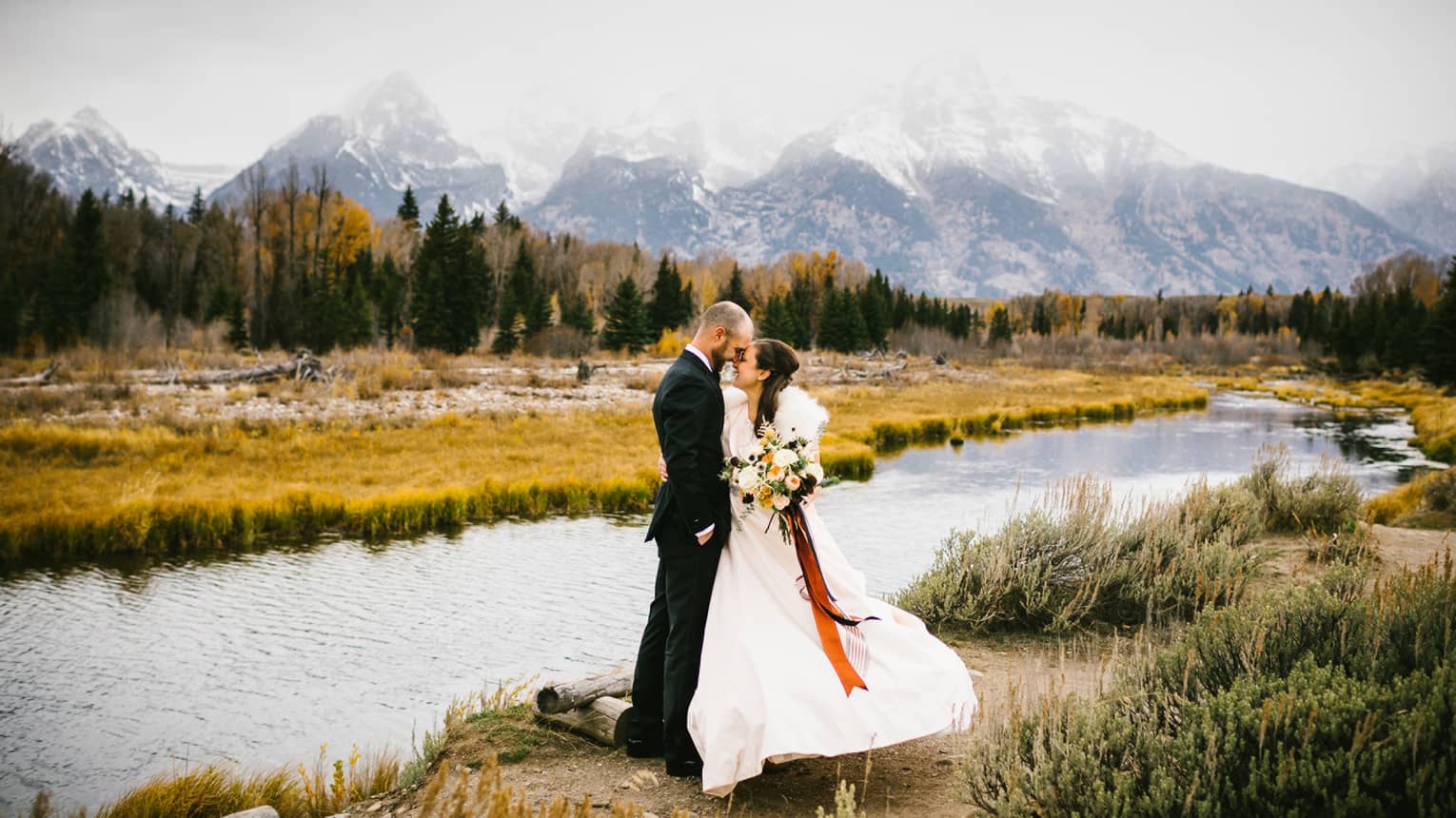 Man in suit and woman in wedding dress with bouquet embrace outdoors with backdrop of river and mountains