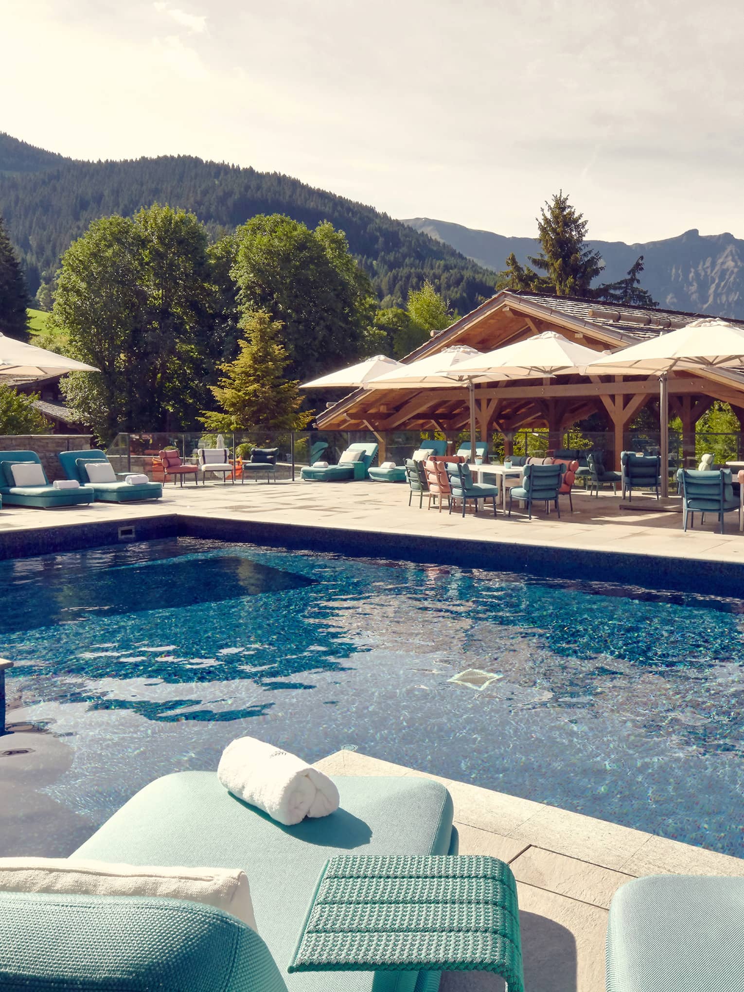 Outdoor pool with aqua blue chaises longues and beige umbrellas on surrounding deck and mountains in background