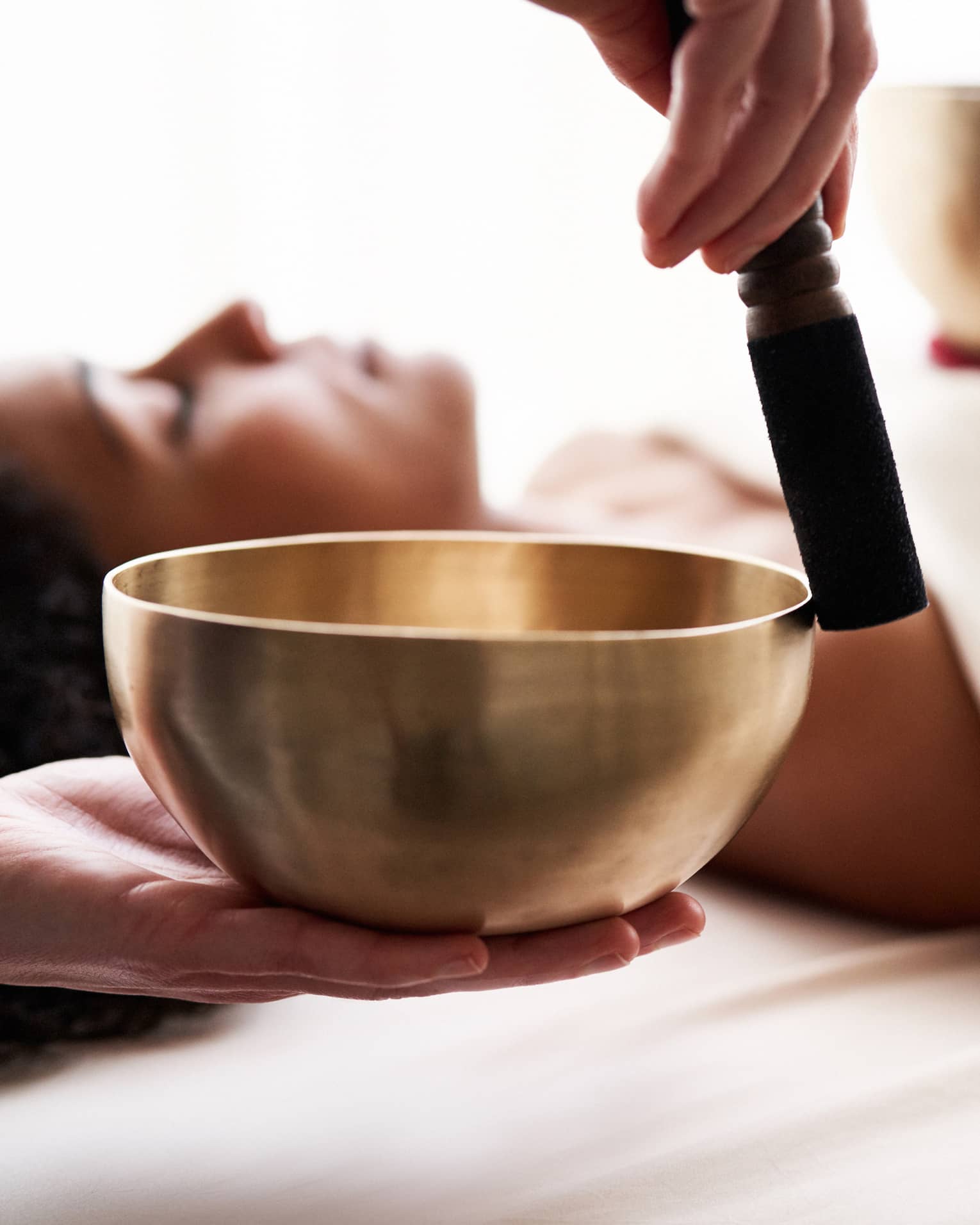 Close up of hands playing a singing bowl with a woman laying down on a spa treatment table in the background