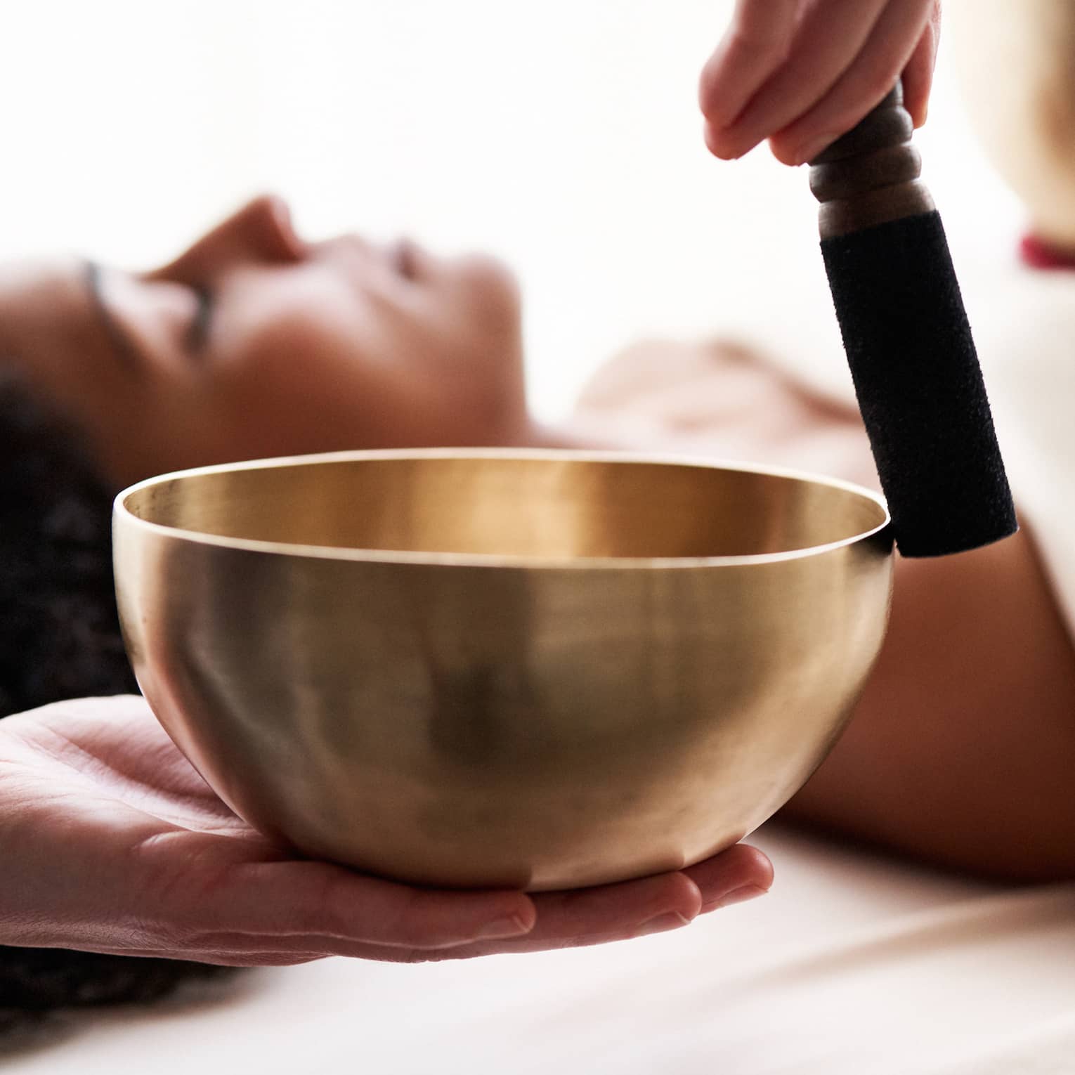 Close up of hands playing a singing bowl with a woman laying down on a spa treatment table in the background