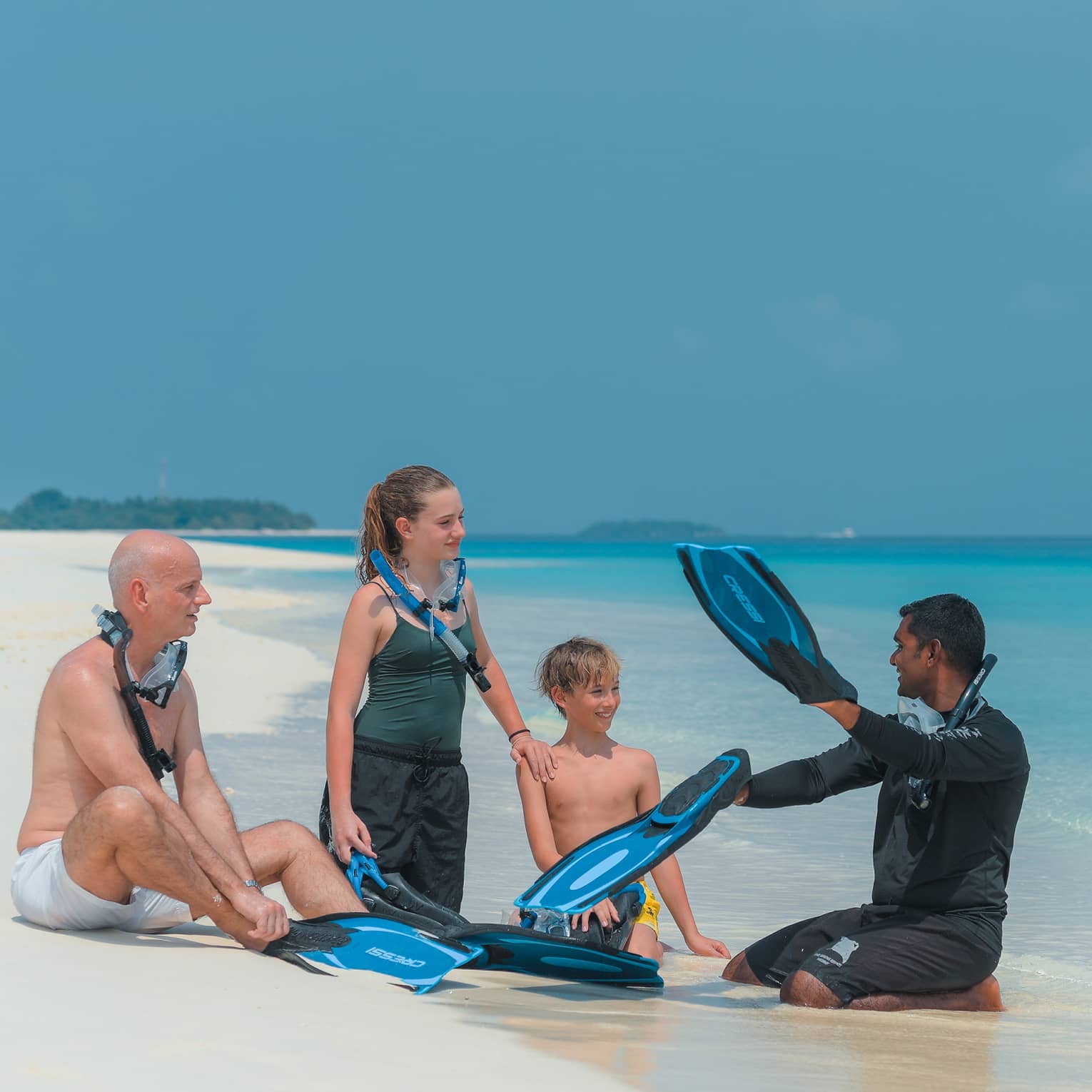 Sitting on a white sandy beach by the water, a family with snorkel gear intently listens to the instructor holding flippers.