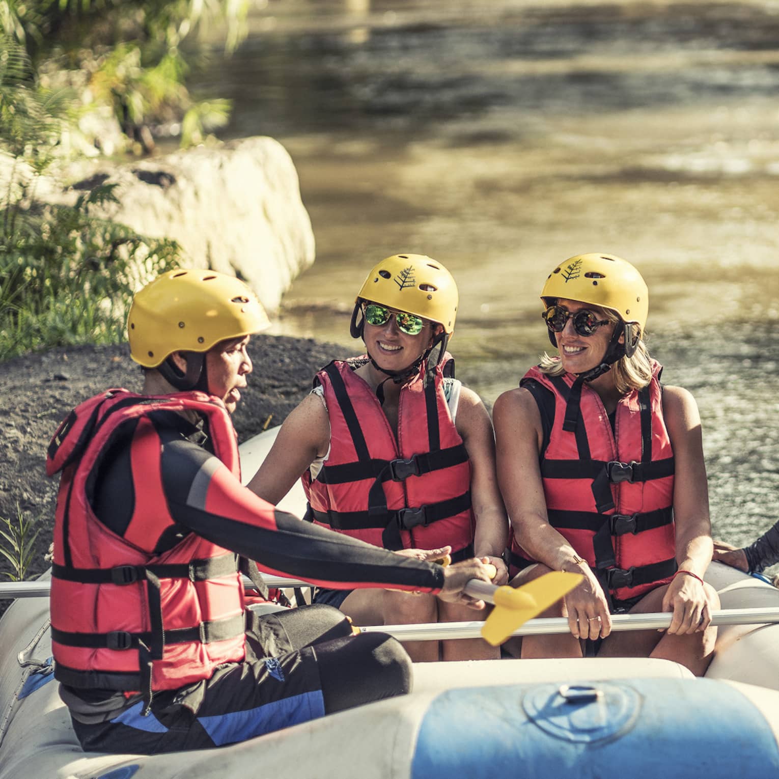 Four people wearing red life jackets, sunglasses and yellow helmets sit facing each other in a river raft