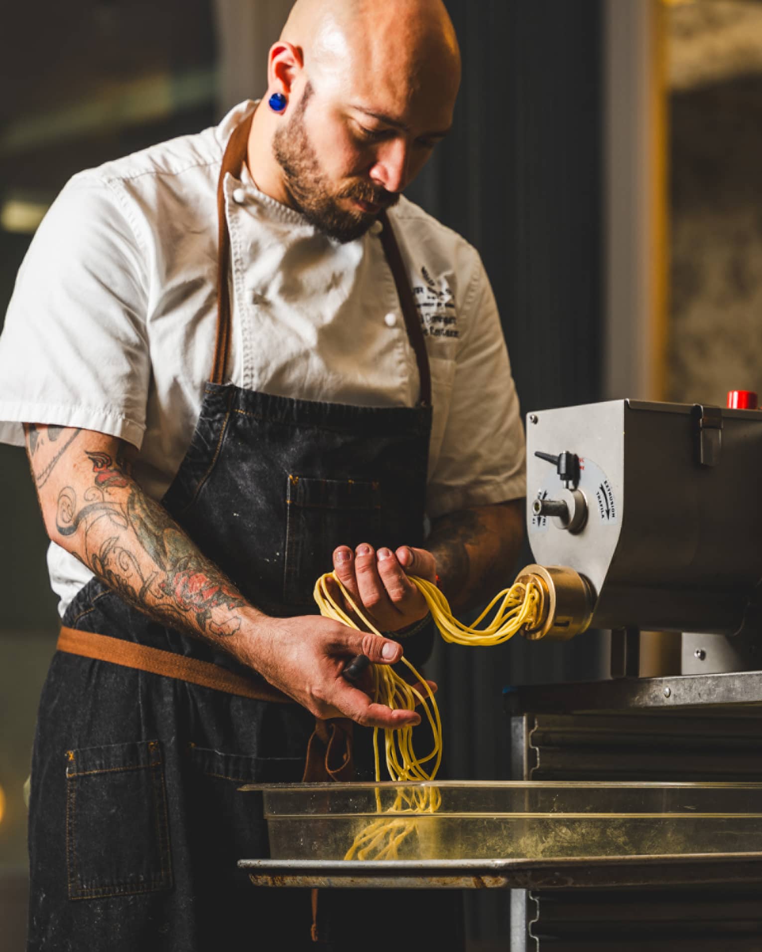 A chef catches fresh long pasta noodles as they emerge from an industrial pasta-making machine in a warmly lit kitchen.