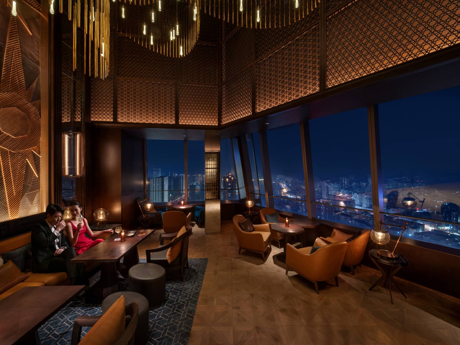 A couple enjoys drinks in a darkly lit bar with ornate lighted ceiling panelling and an aerial view of the city at night.