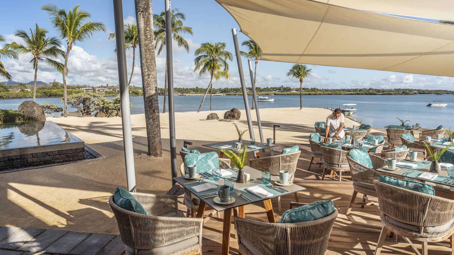 Server setting tables flanked with bamboo chairs and aqua pillows under tan shade at waterfront 