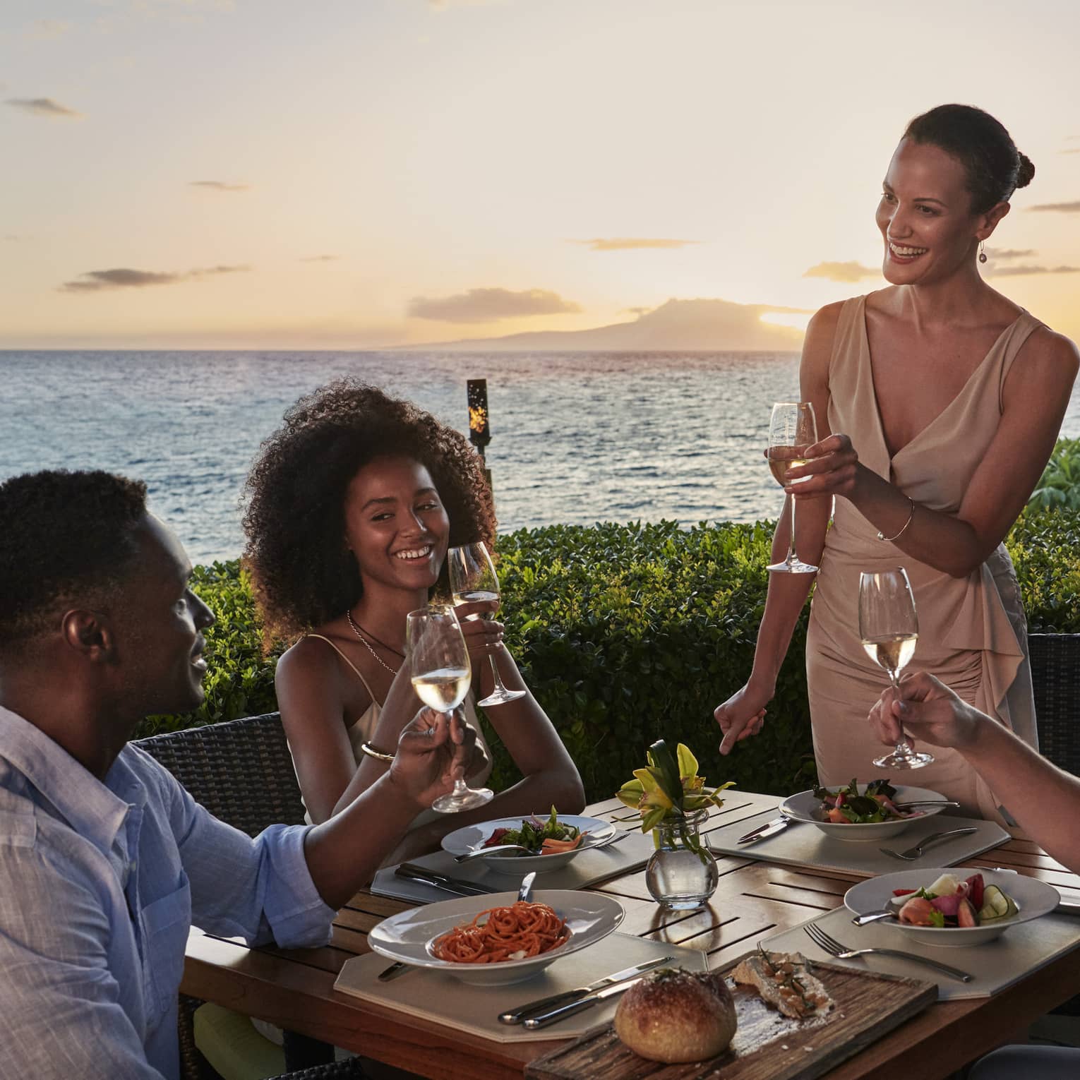 Two couples dine outside at Ferraro’s Bar E Ristorante, overlooking Wailea Beach. The sun is setting as they raise a toast. 